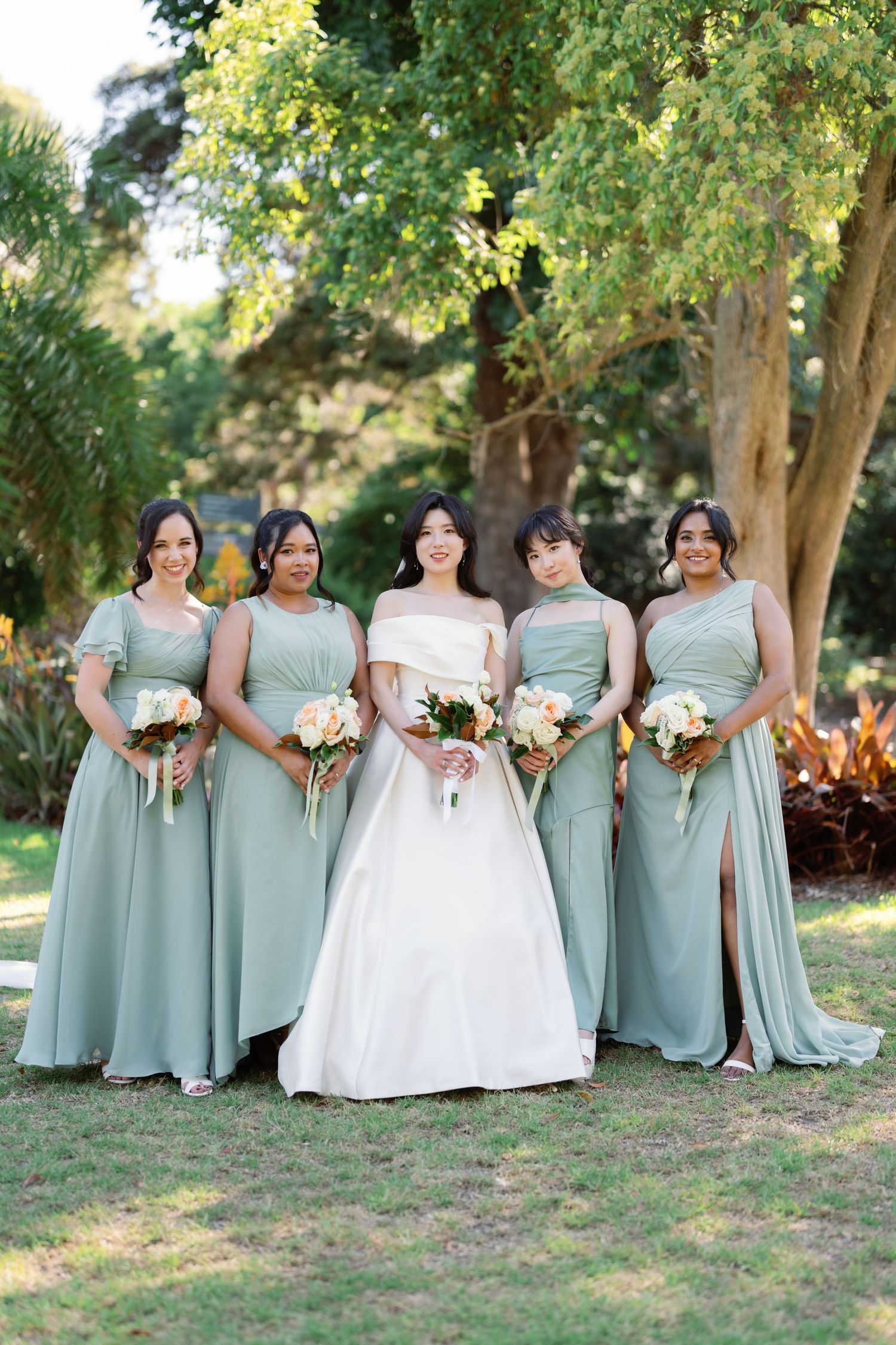 Bridal party in sage green dresses and white wedding gown pose together on grassy hill.