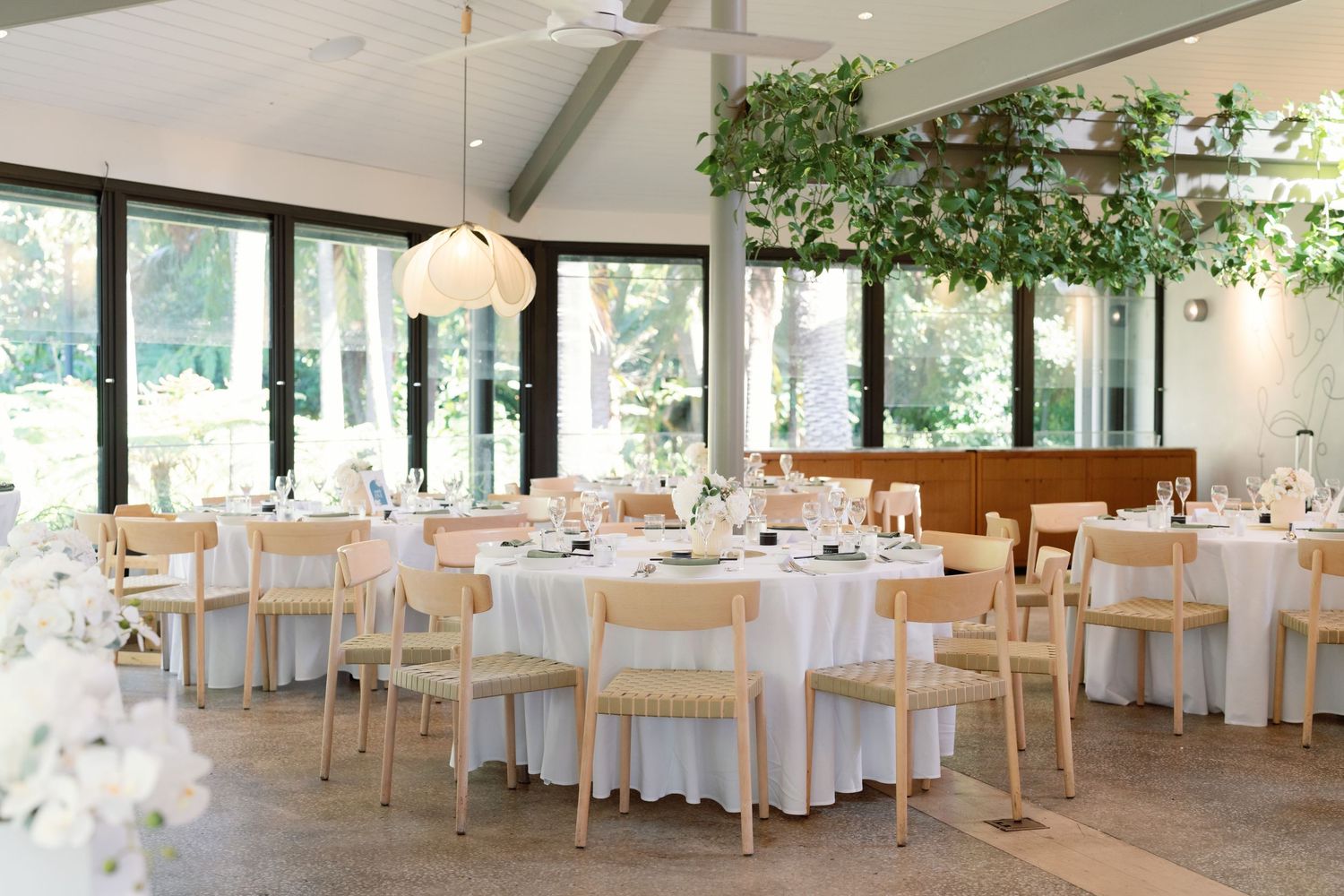 Elegant wedding reception venue with round tables, white linens, wooden chairs and natural light from floor-to-ceiling windows.