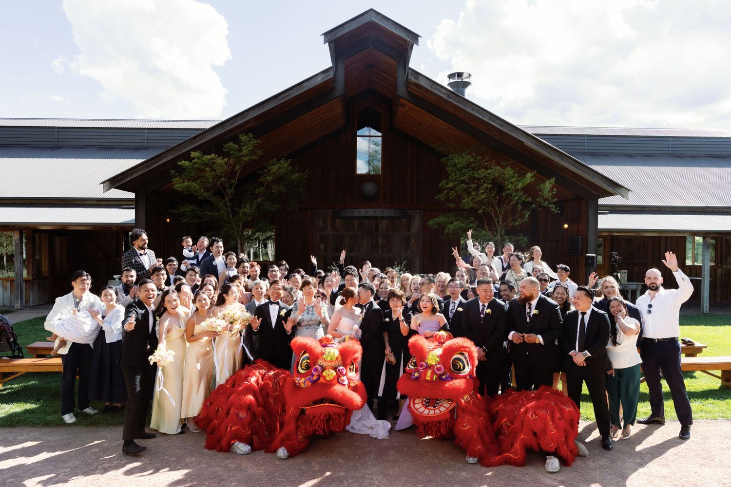 Large wedding group poses with red Chinese lion dance performers outside a wooden venue.