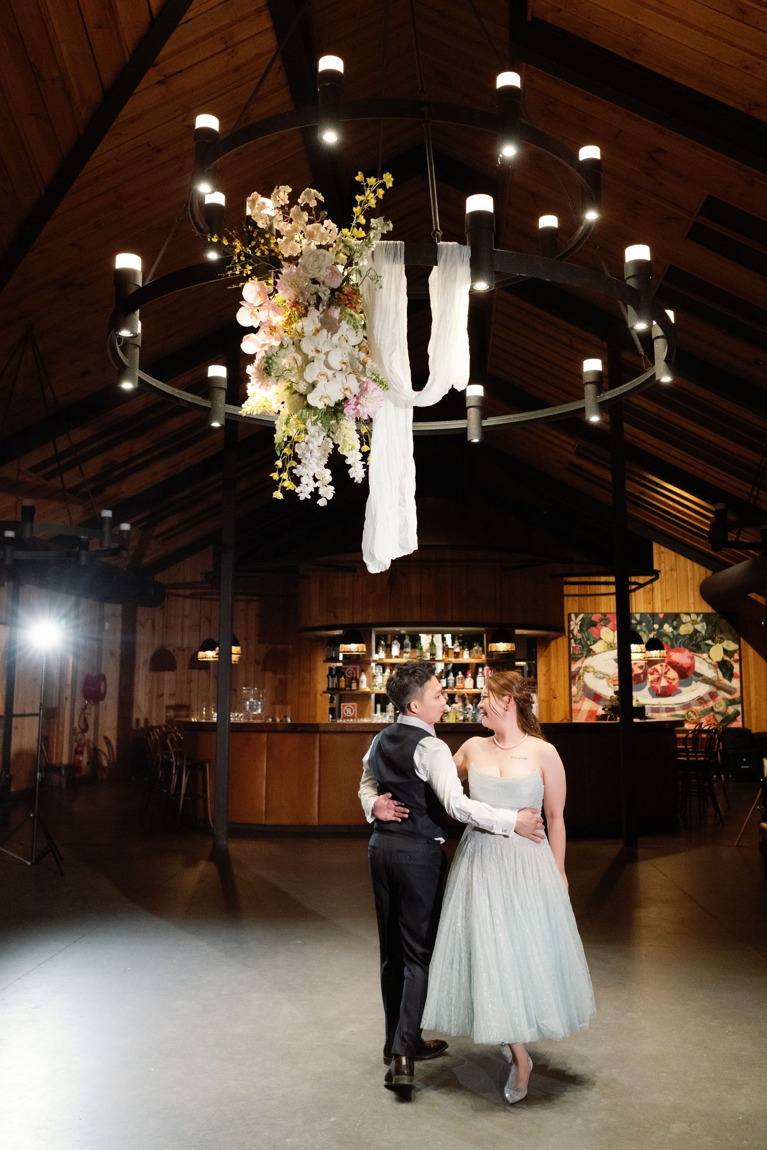 A couple stands beneath an elegant chandelier decorated with flowers and fabric in a rustic wooden venue.
