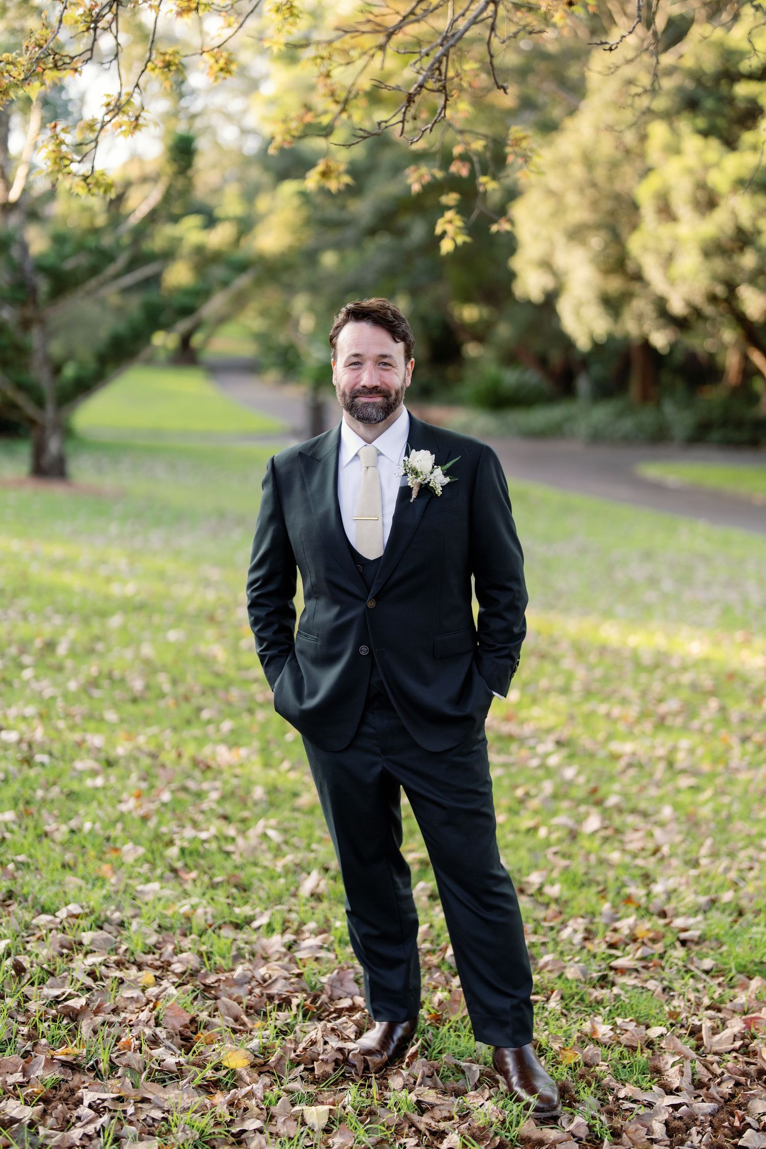 A bearded groom in a dark suit with floral boutonniere stands confidently among autumn leaves in a park.