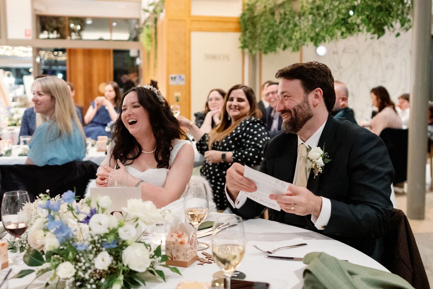 Bride and groom laughing joyfully during wedding reception speeches at elegantly decorated indoor venue.
