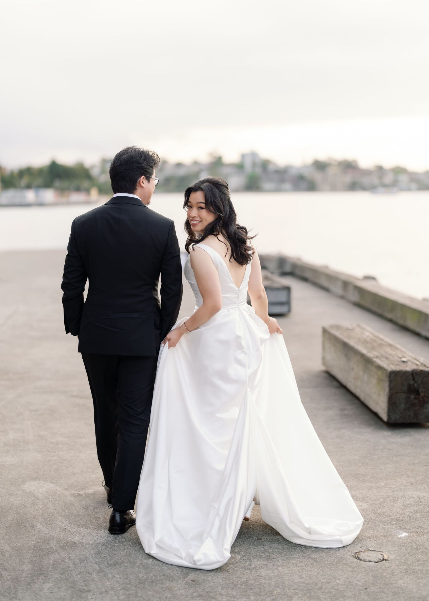 Couple in wedding attire walking along waterfront promenade.