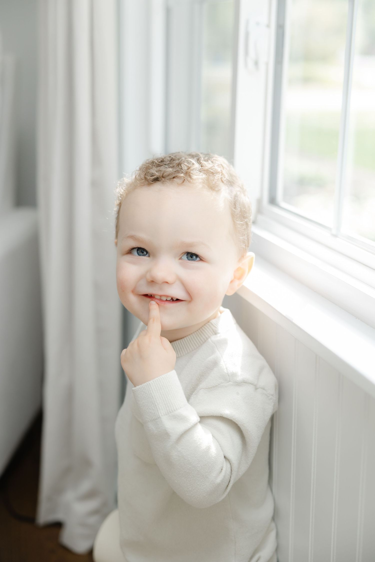 A baby wearing a white sweater smiles while looking out a bright window.