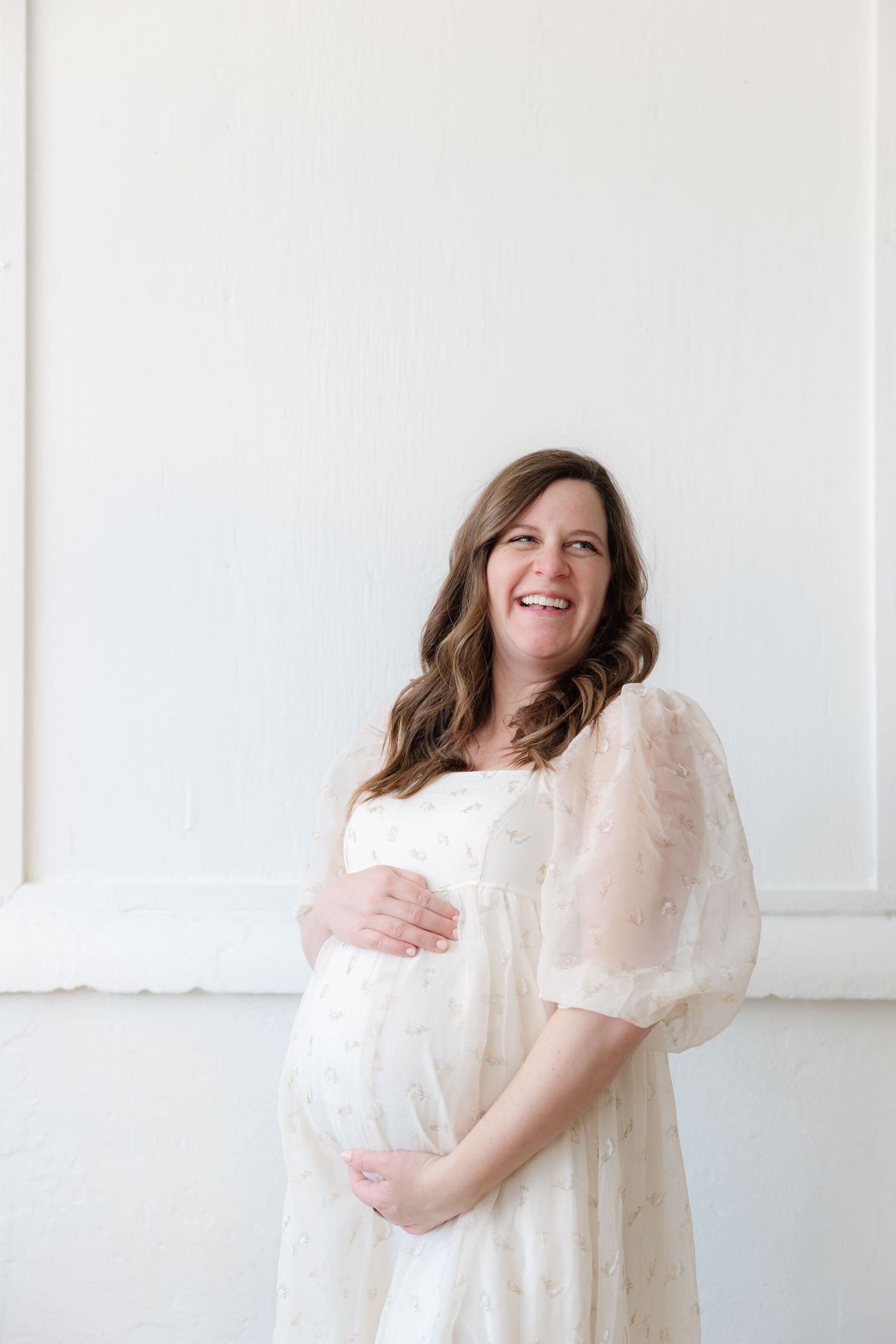 A pregnant woman in a flowing white dress smiles while standing against a white wall in a bright minimalist setting.