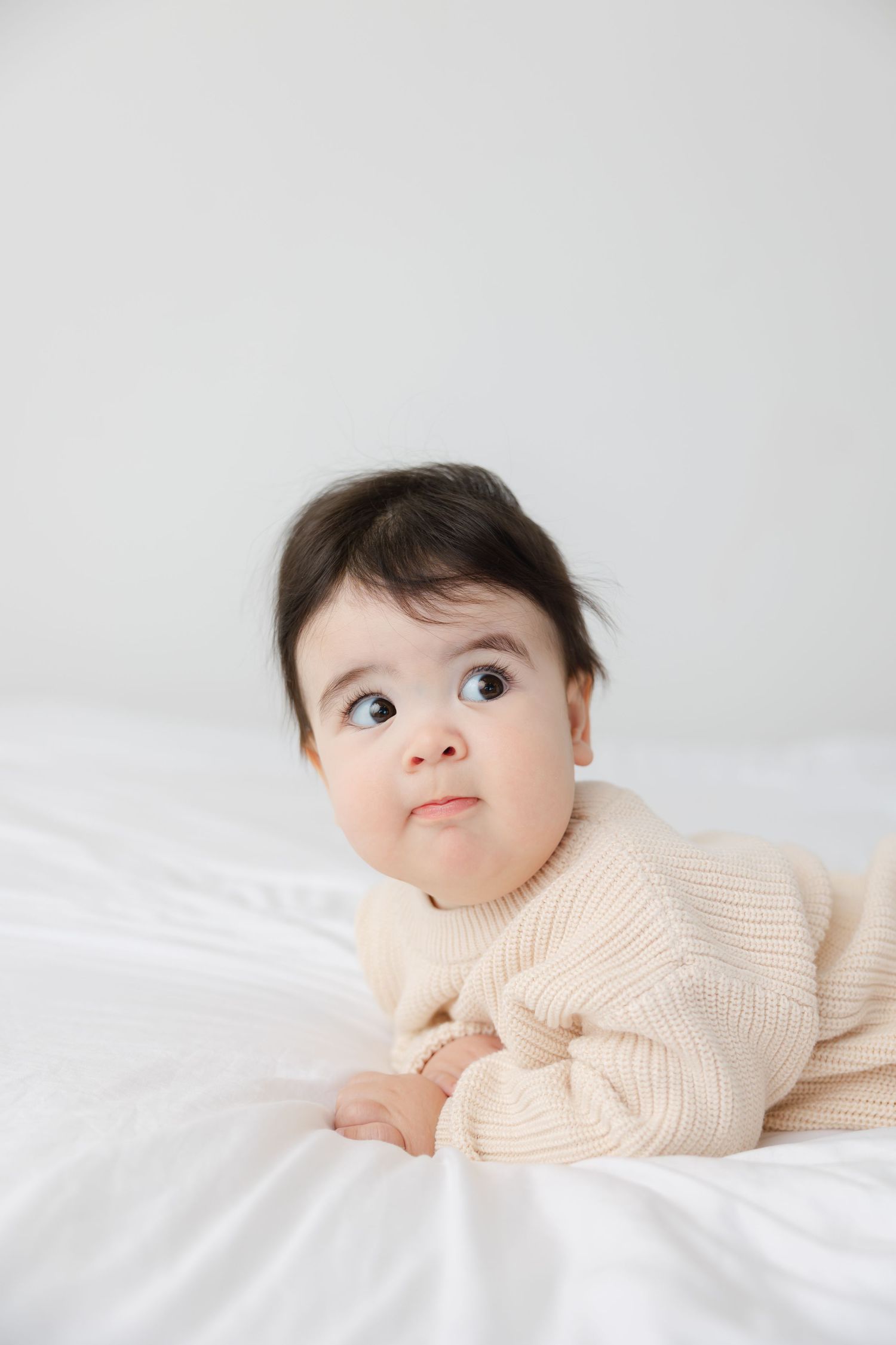 Baby in cream knit sweater lying on white bedding looking upward with curious expression.