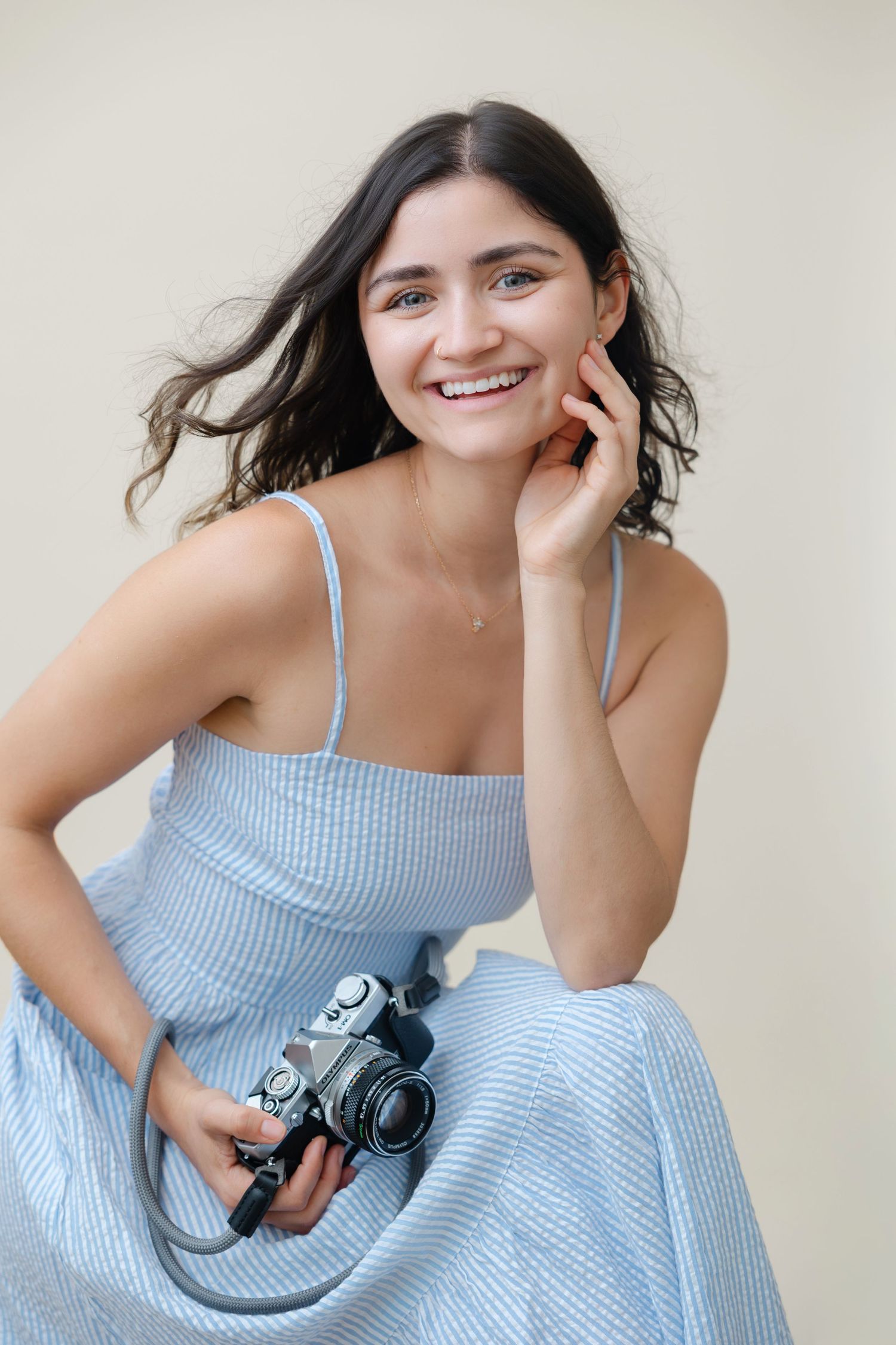 Woman in light blue summer dress smiling while holding vintage camera against neutral background.