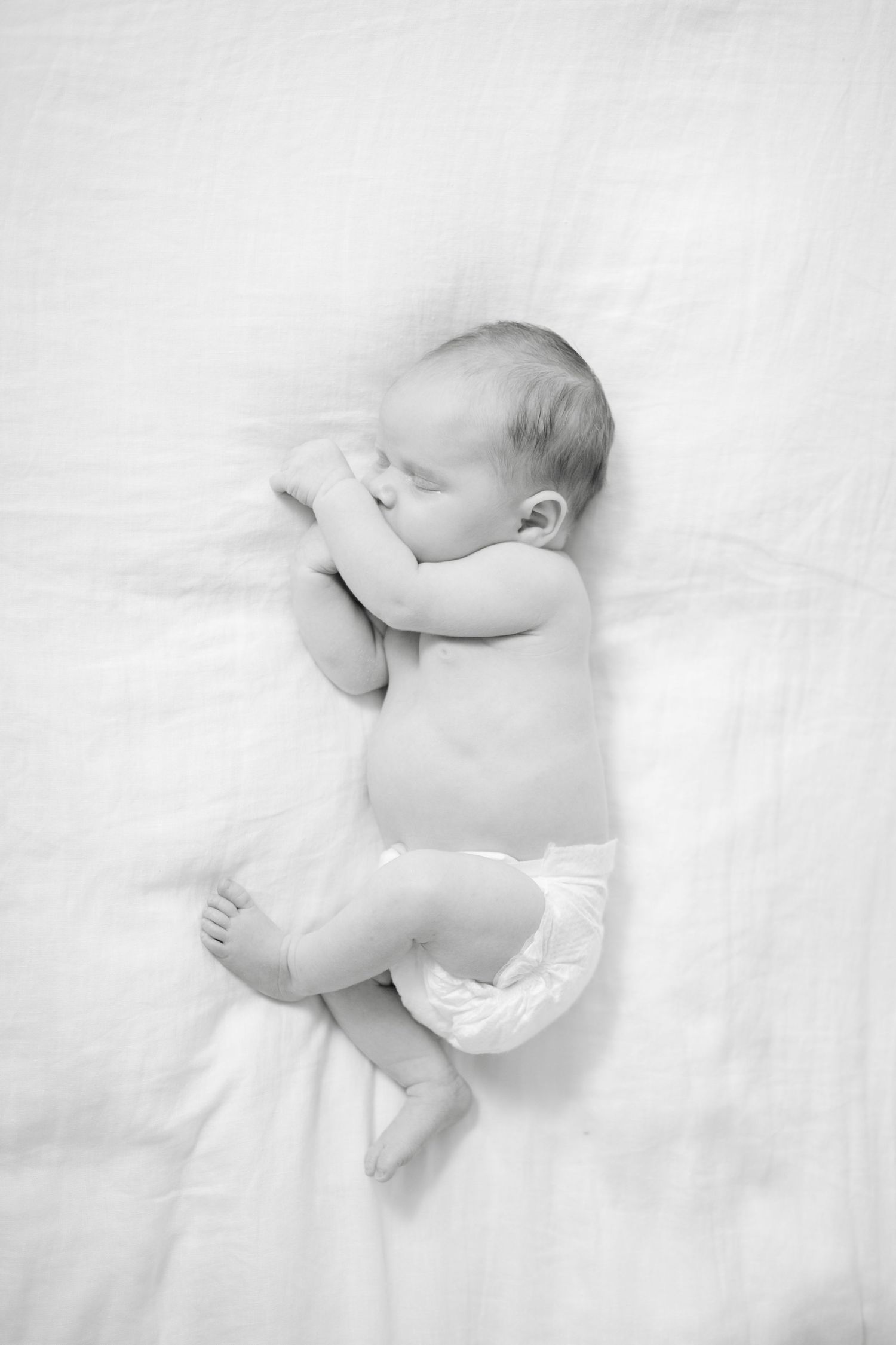 High key black and white portraits of a sleeping newborn baby wearing white against white bedding.