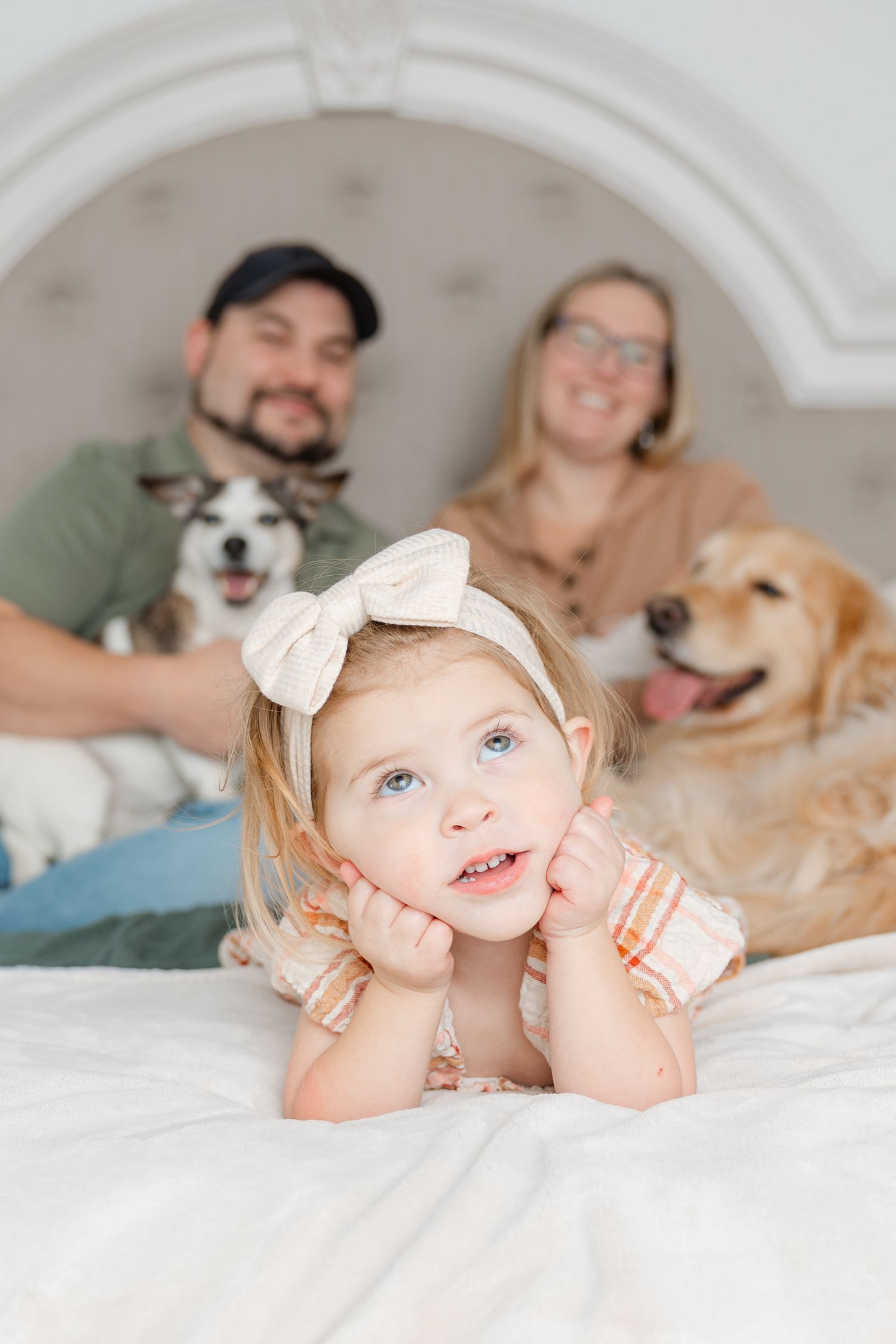 A heartwarming family portrait on white bedding with a toddler in a bow and two dogs in the background.