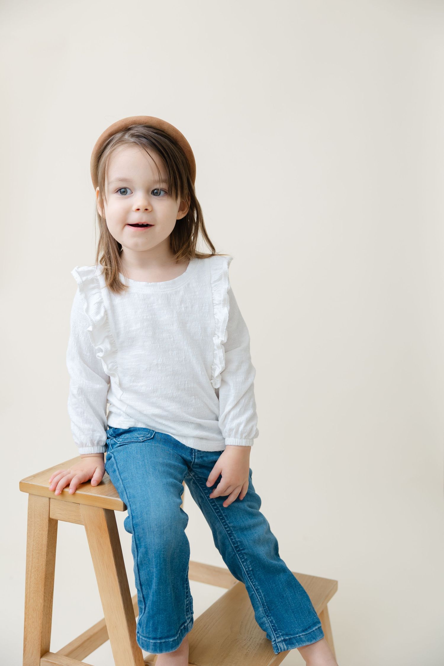 A young child in a white sweater and jeans sits casually on a wooden stool against a light background.