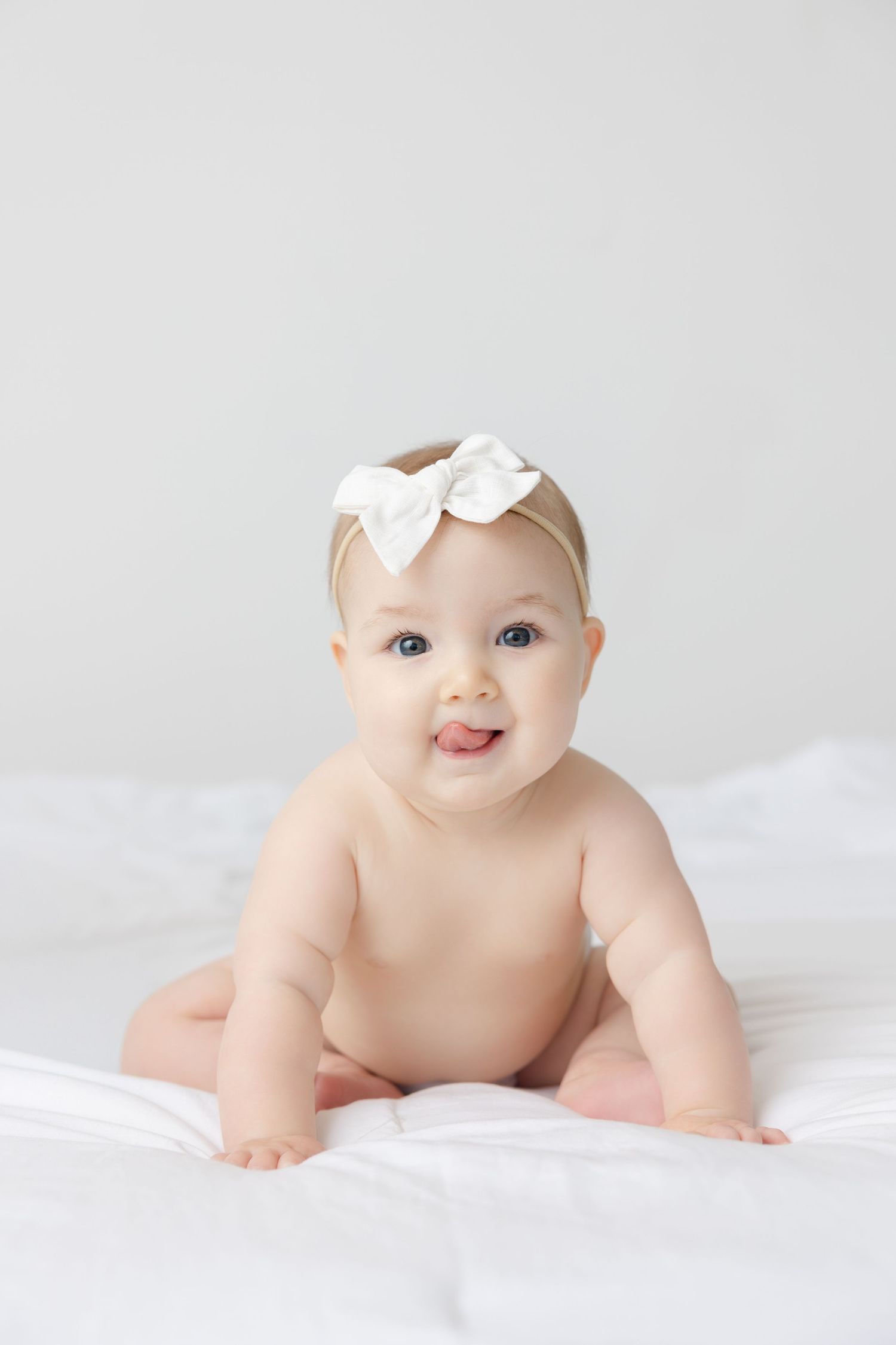 Sweet baby wearing white bow crawls on soft white blanket in bright minimalist portrait series.