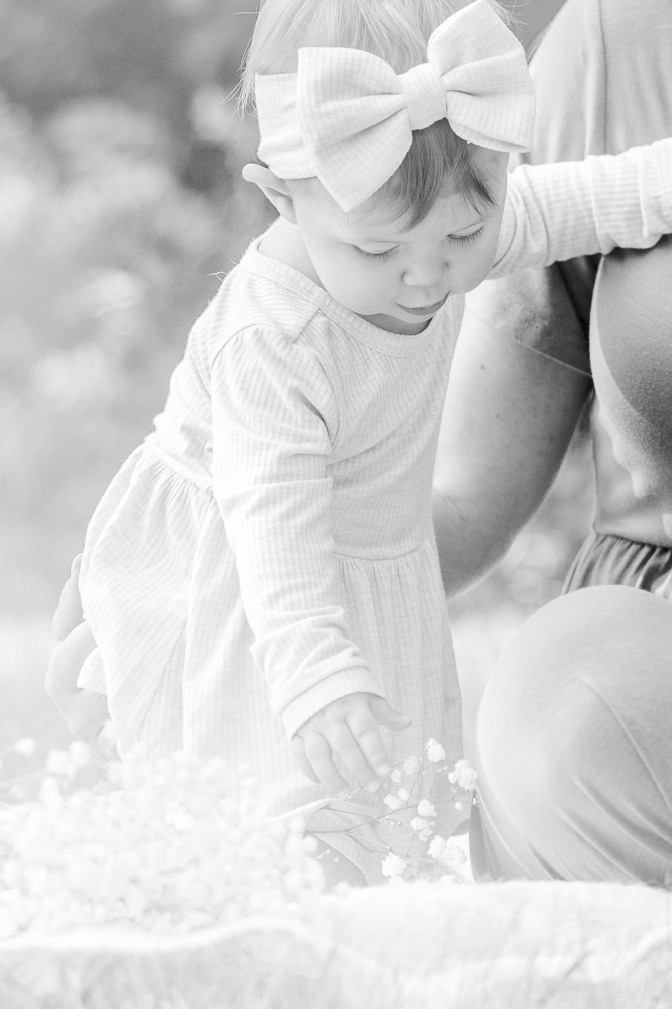 A small child in a white shirt bends down to examine flowers in a soft, dreamy black and white photograph.