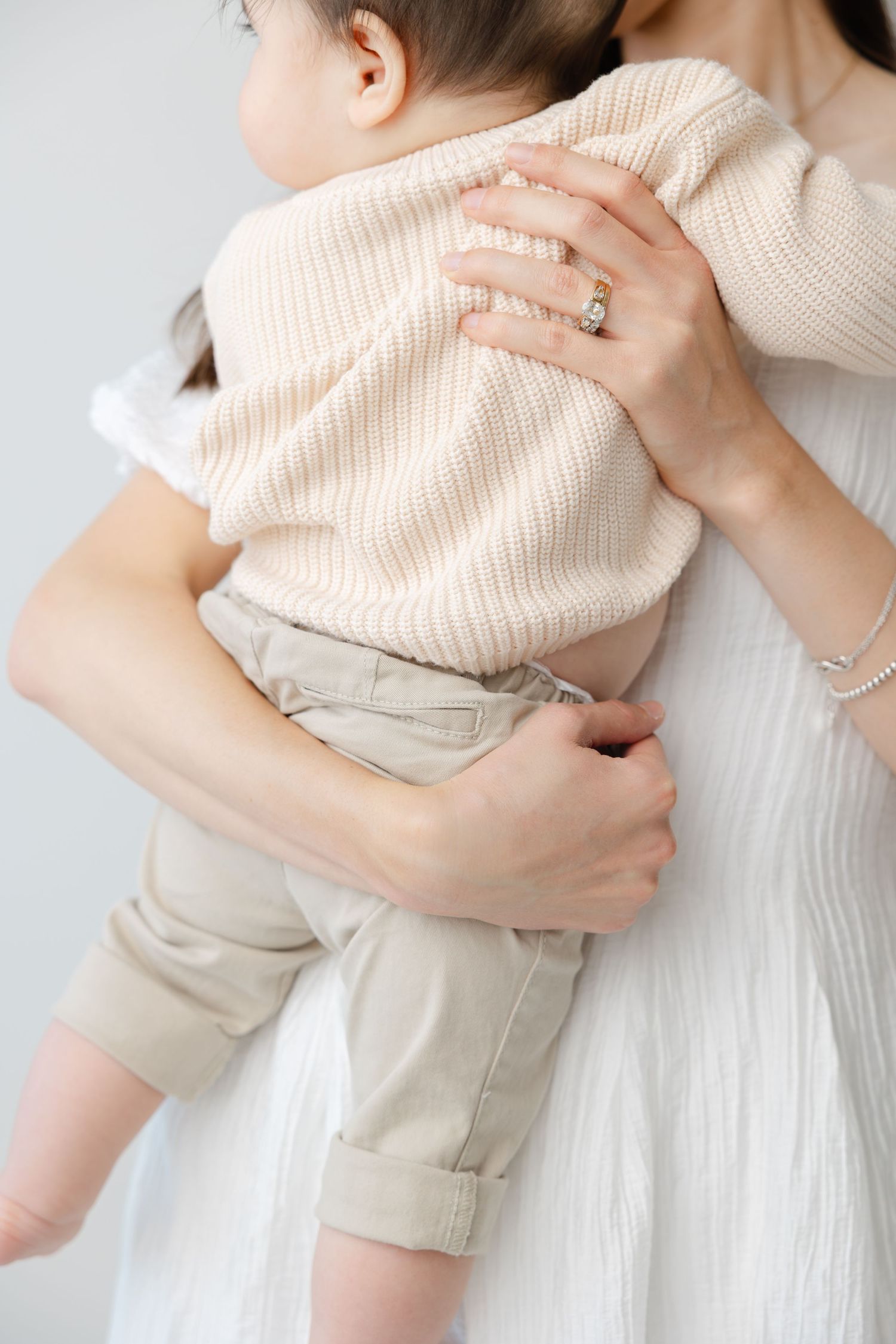 A parent in a white dress holds a baby wearing a beige sweater in a series of tender embraces.
