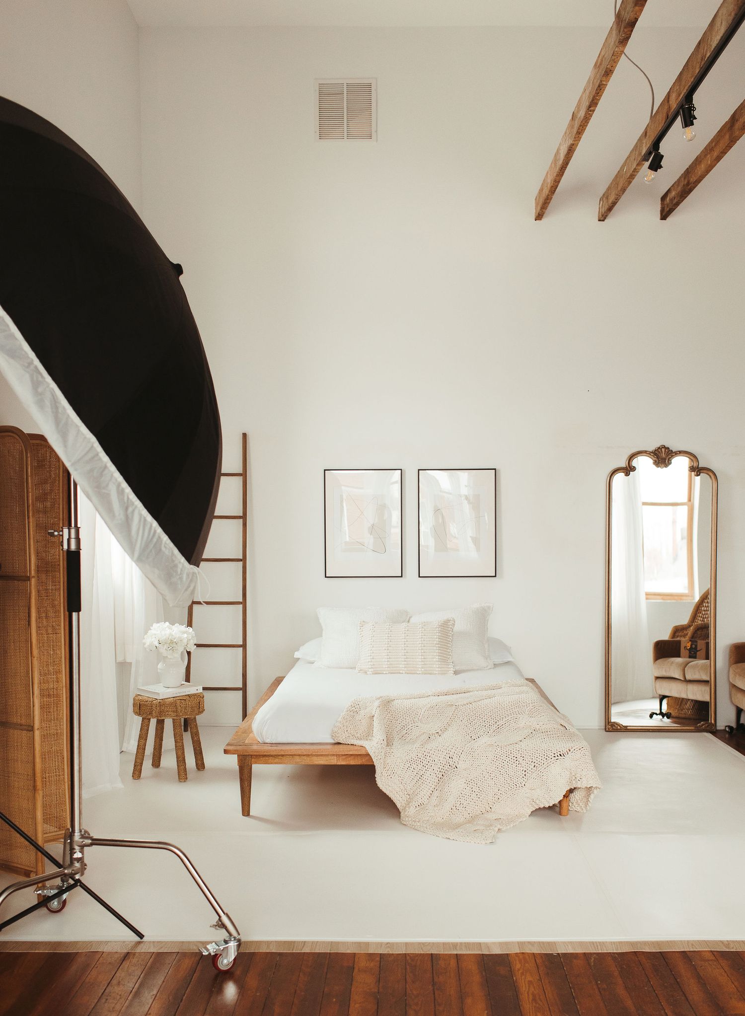 Minimalist bedroom with white walls, wooden beams, vintage mirror, and photography studio equipment in natural light.