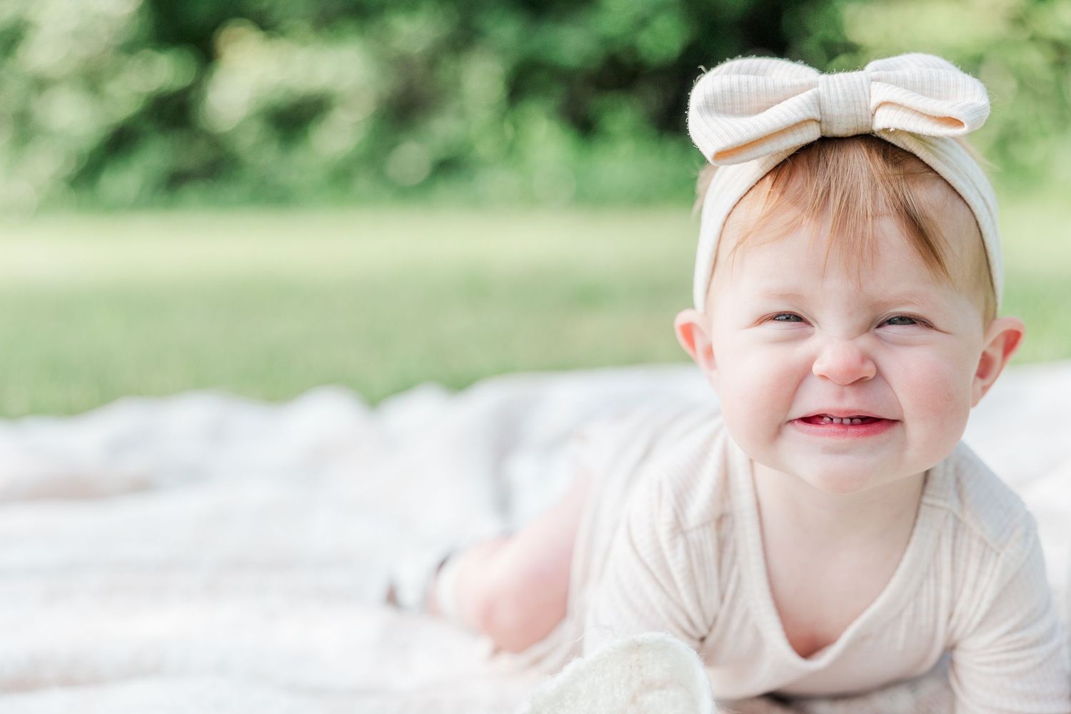 Sweet child wearing light beige clothes and bow headband smiles brightly in outdoor garden setting.