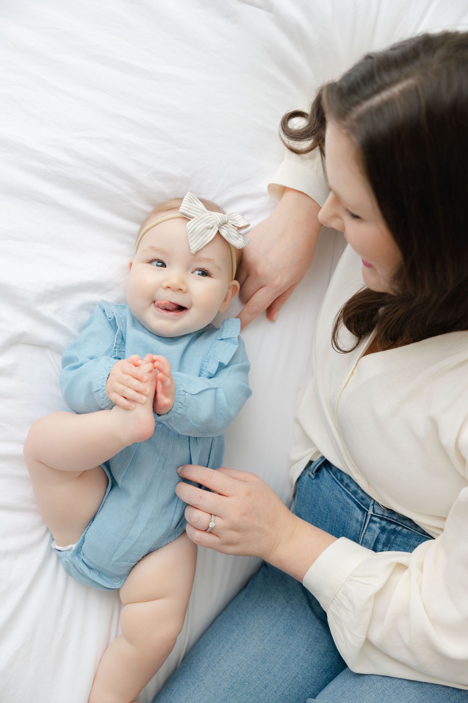 A baby in a blue dress lies on white bedding while a caregiver's hands gently interact during a tender moment.