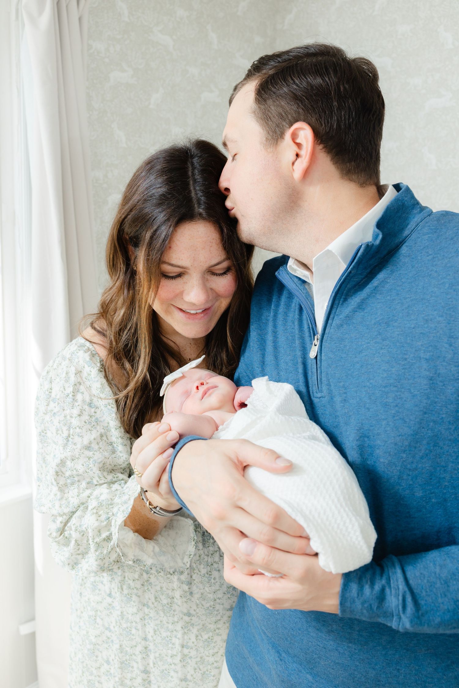 A couple in blue and white clothing holds and gazes lovingly at their newborn baby near a bright window.