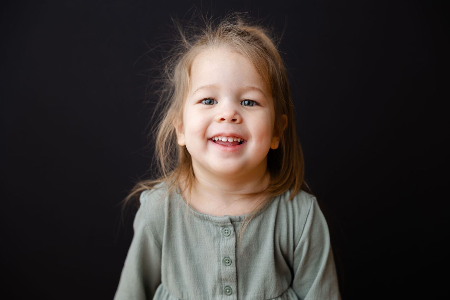 A young child wearing a sage green top smiles brightly against a dark background.