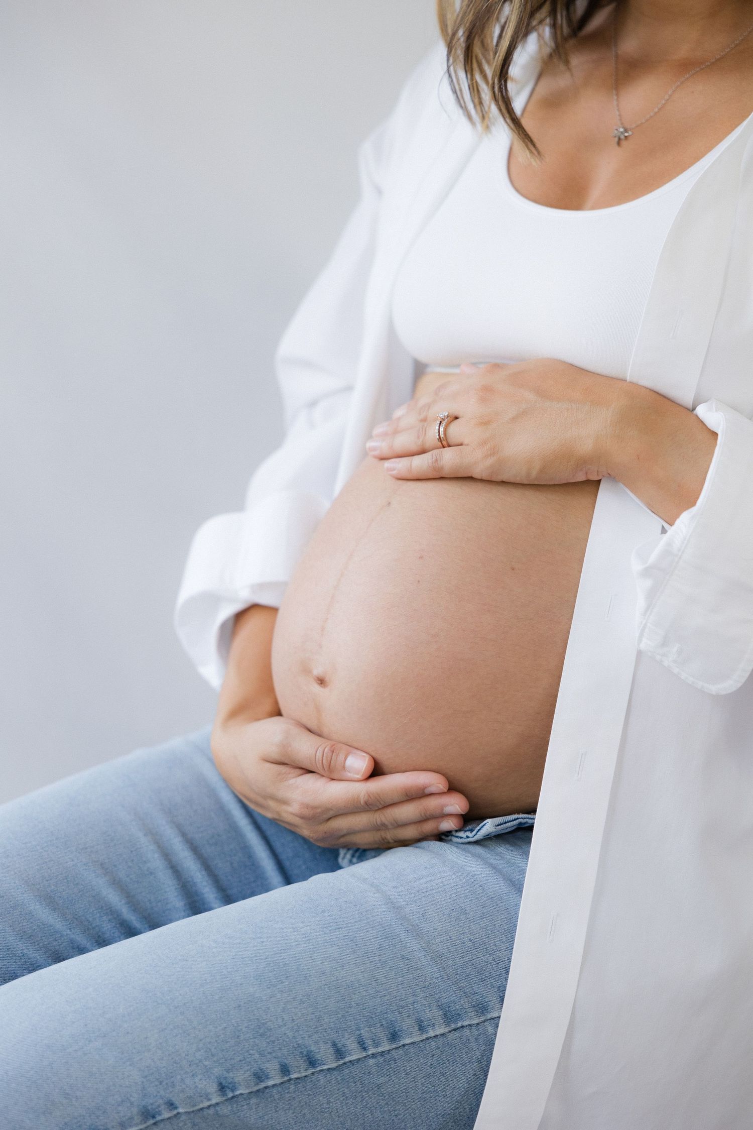 Close-up of pregnant belly with hands gently cradling it, wearing white top and blue jeans.