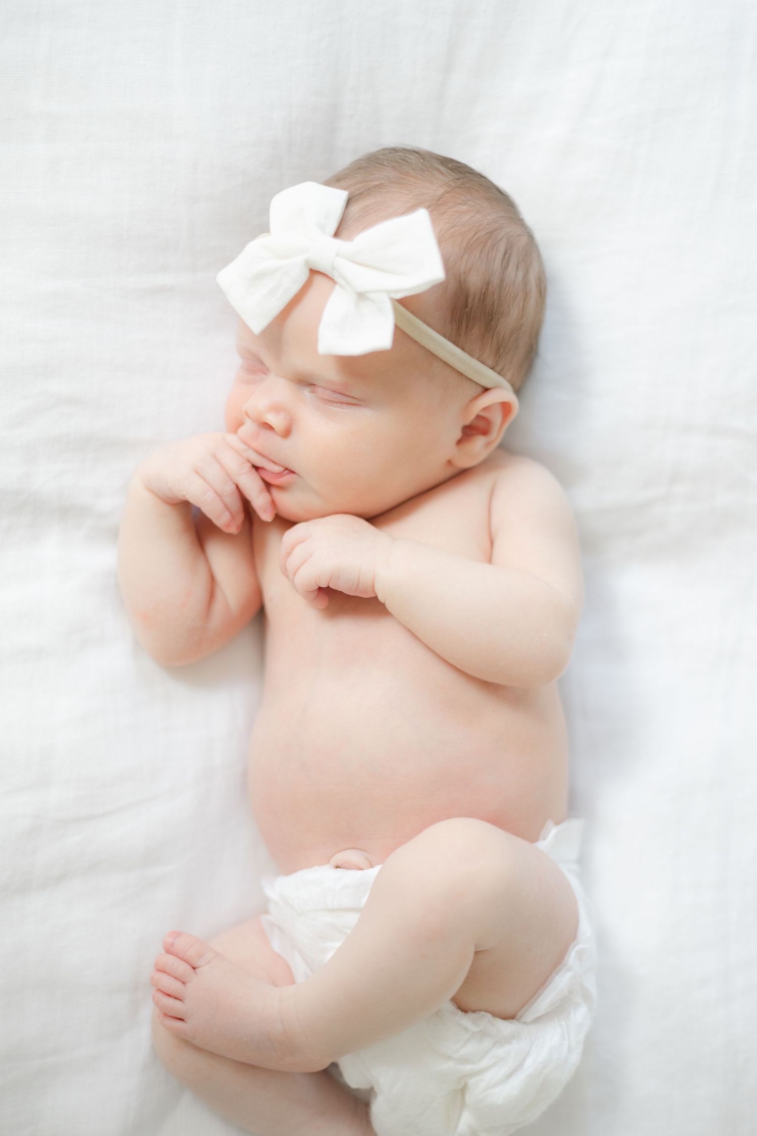 Sleeping newborn baby in white diaper and bow headband lying on soft white bedding.