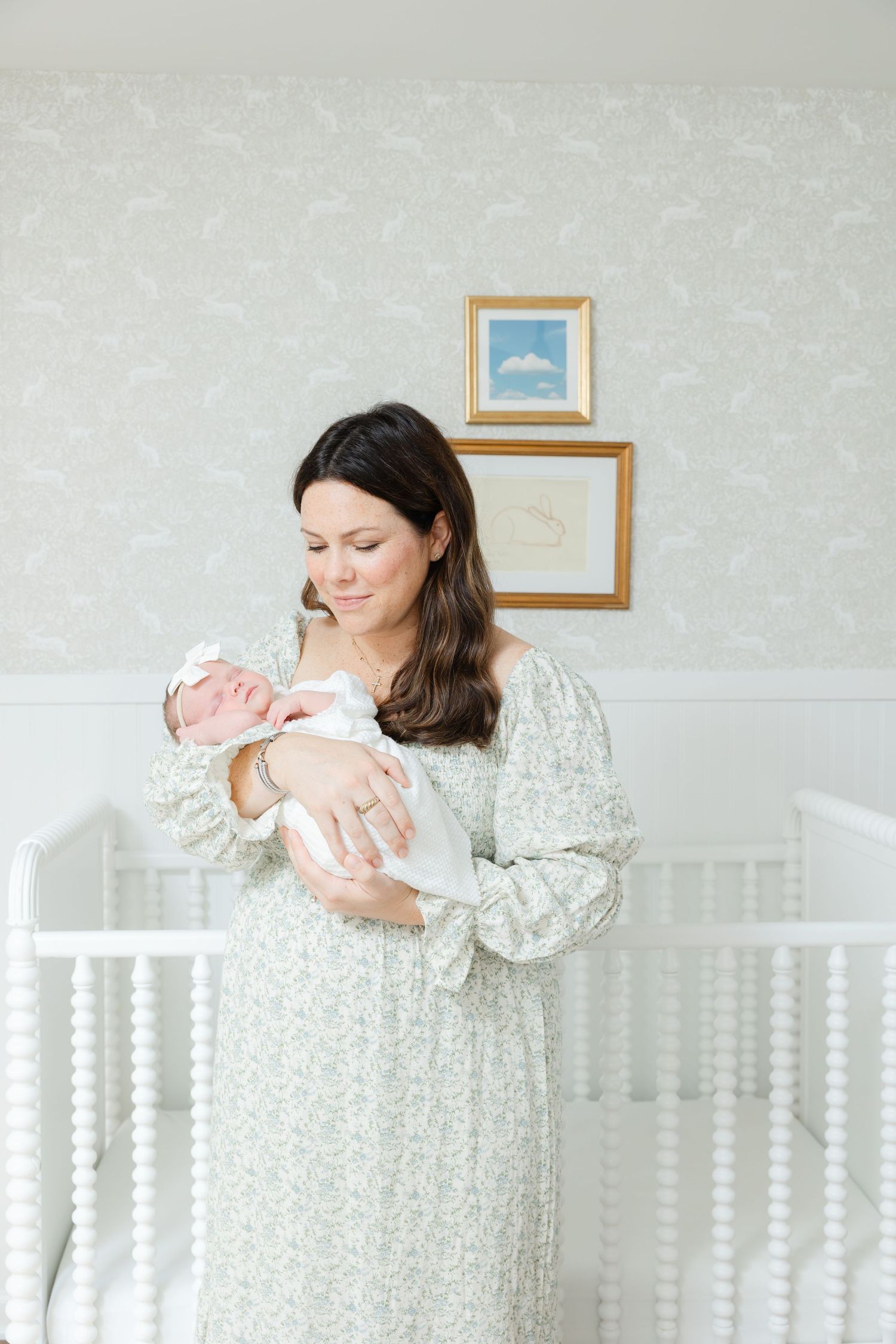 Parent in a floral dress tenderly holds and gazes at their newborn baby in a bright white nursery room.