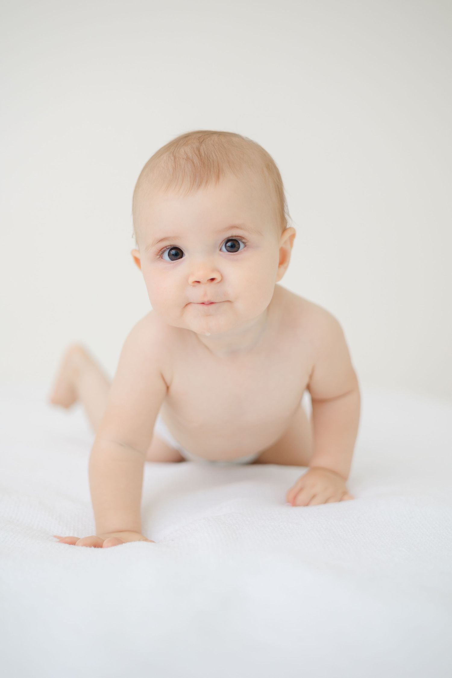A baby in white surroundings crawls and explores while wearing only a diaper.