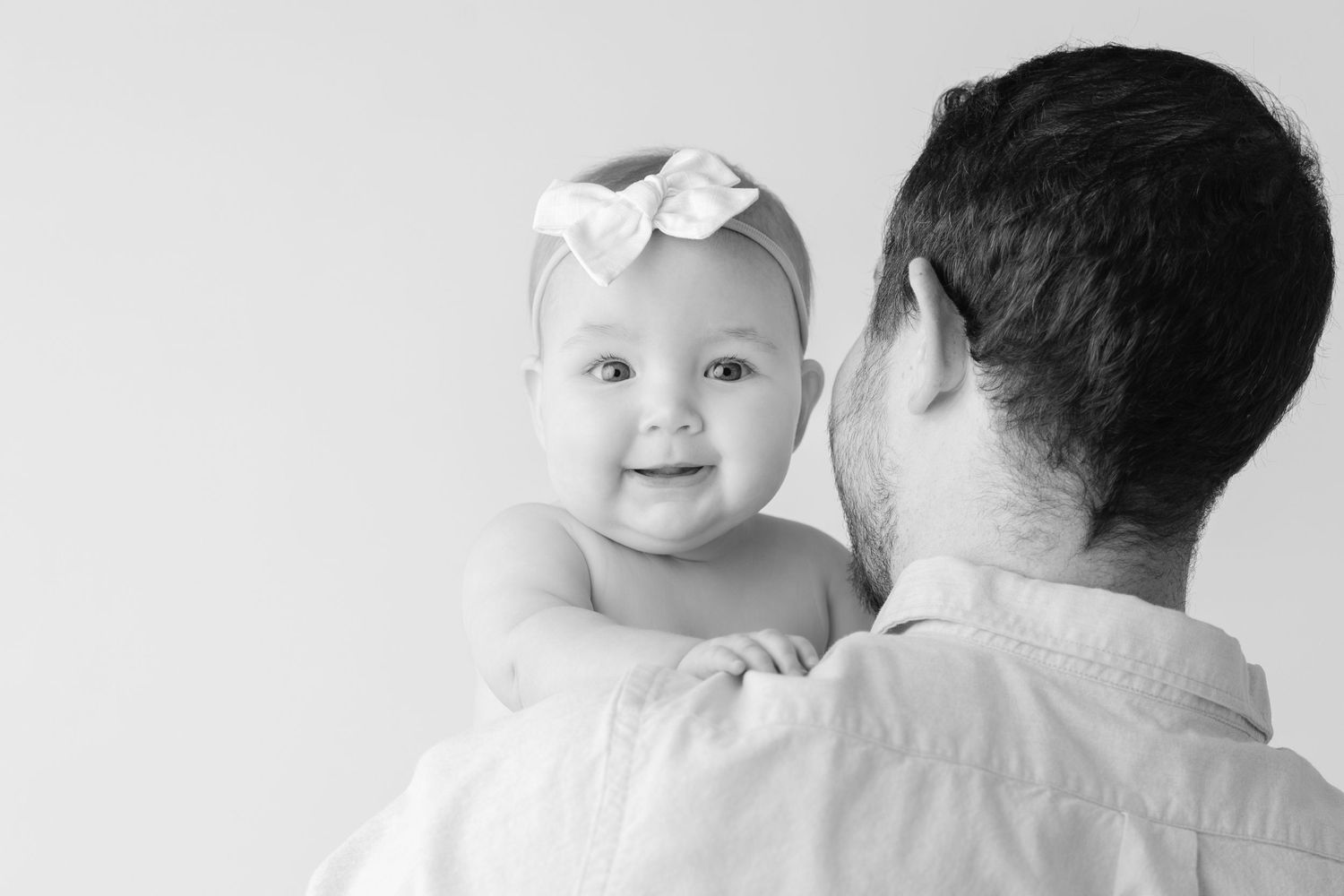 A baby in a white outfit smiles while being held by someone wearing a black shirt.