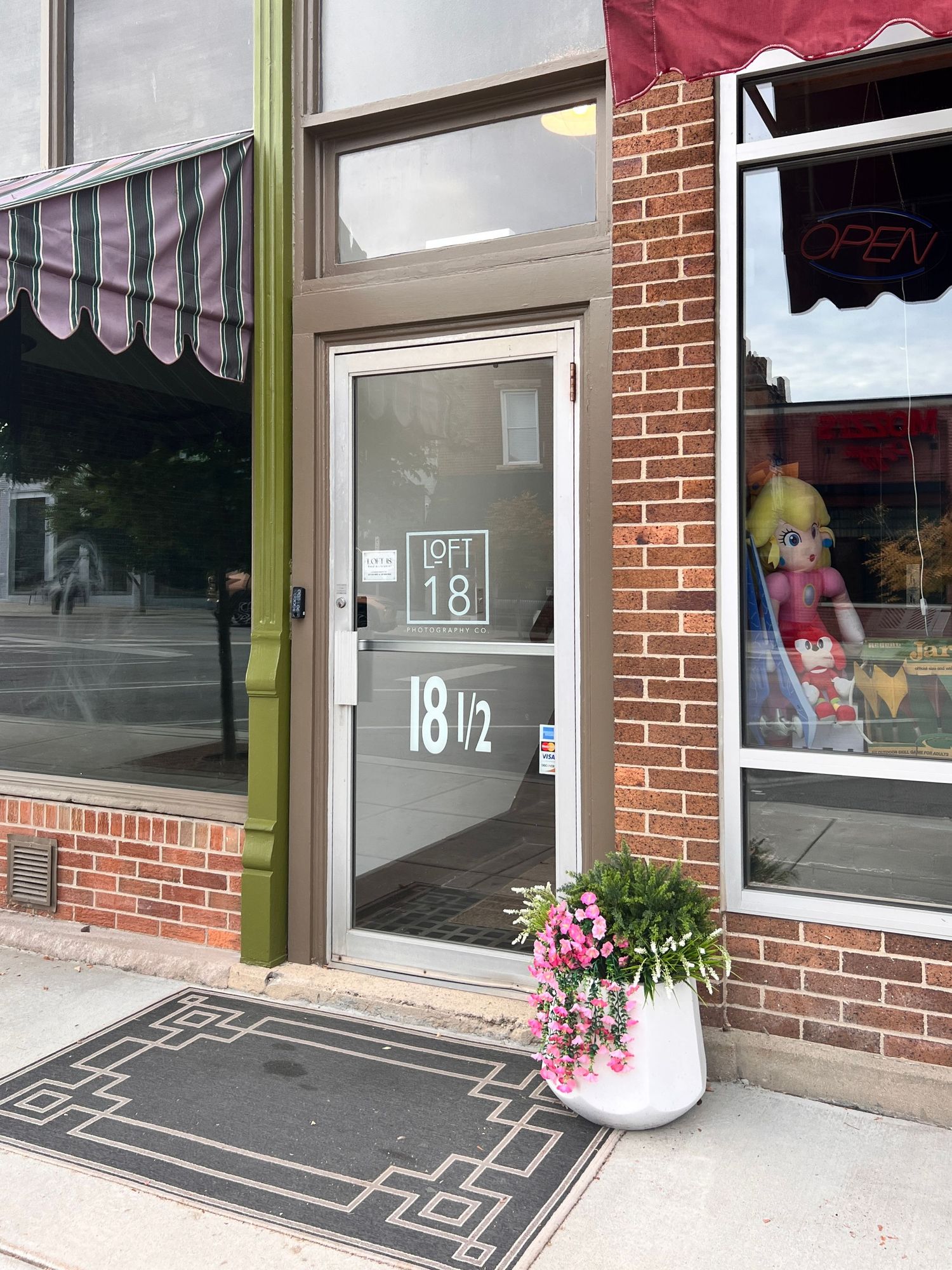 A series of storefront windows with decorative doormats and pink flowers in planters along a brick building exterior.