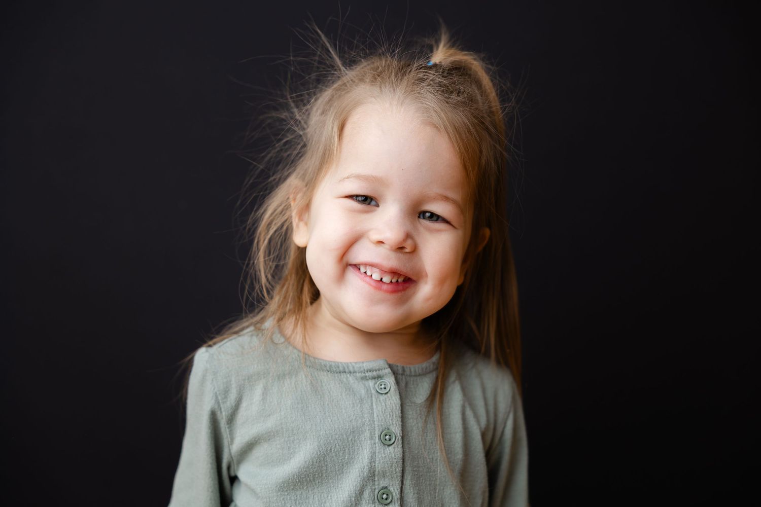 Child in sage green cardigan smiles brightly against dark background.