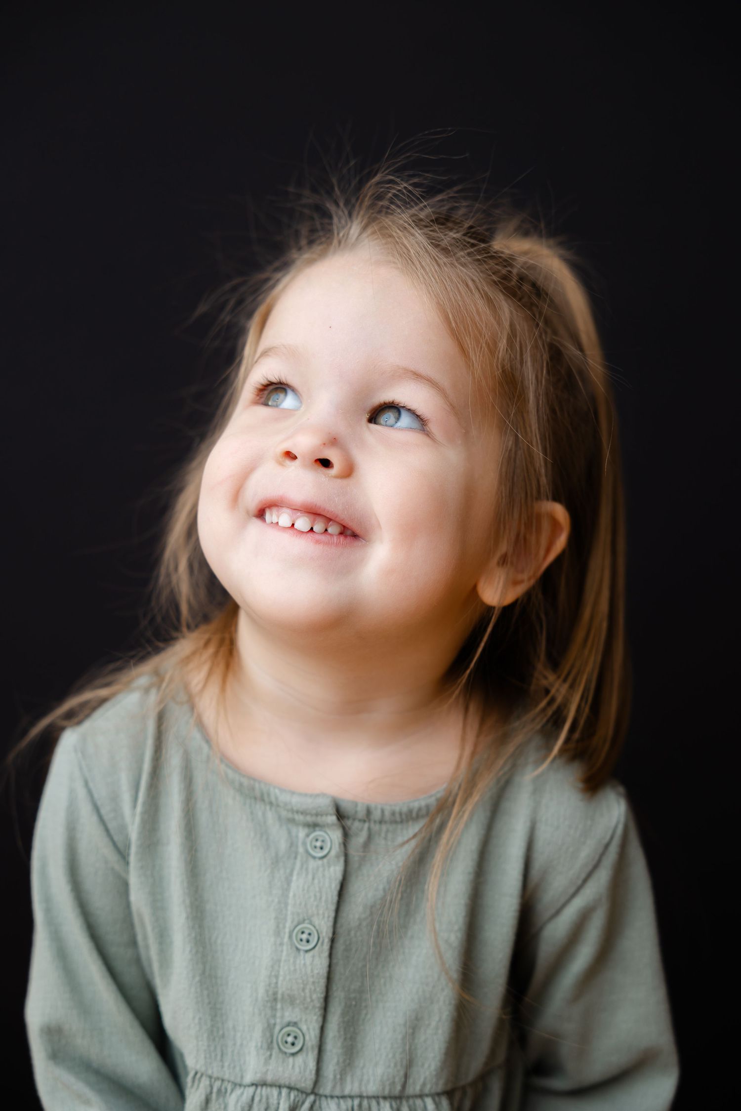 Young child in sage green dress smiles and gazes upward against dark background in heartwarming portrait.
