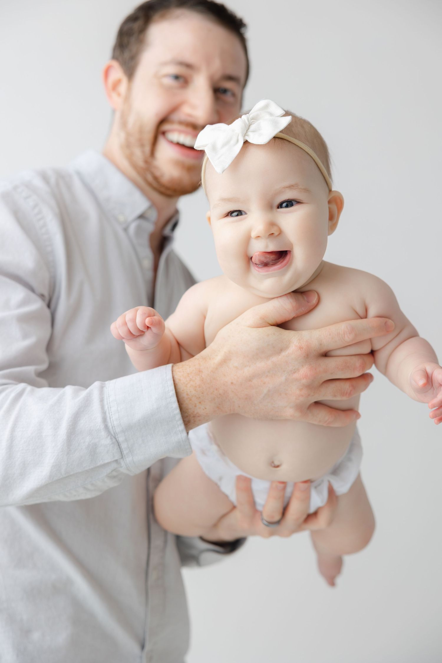 A baby wearing a white bow laughs while being held by someone in a light gray shirt in a series of four photos.