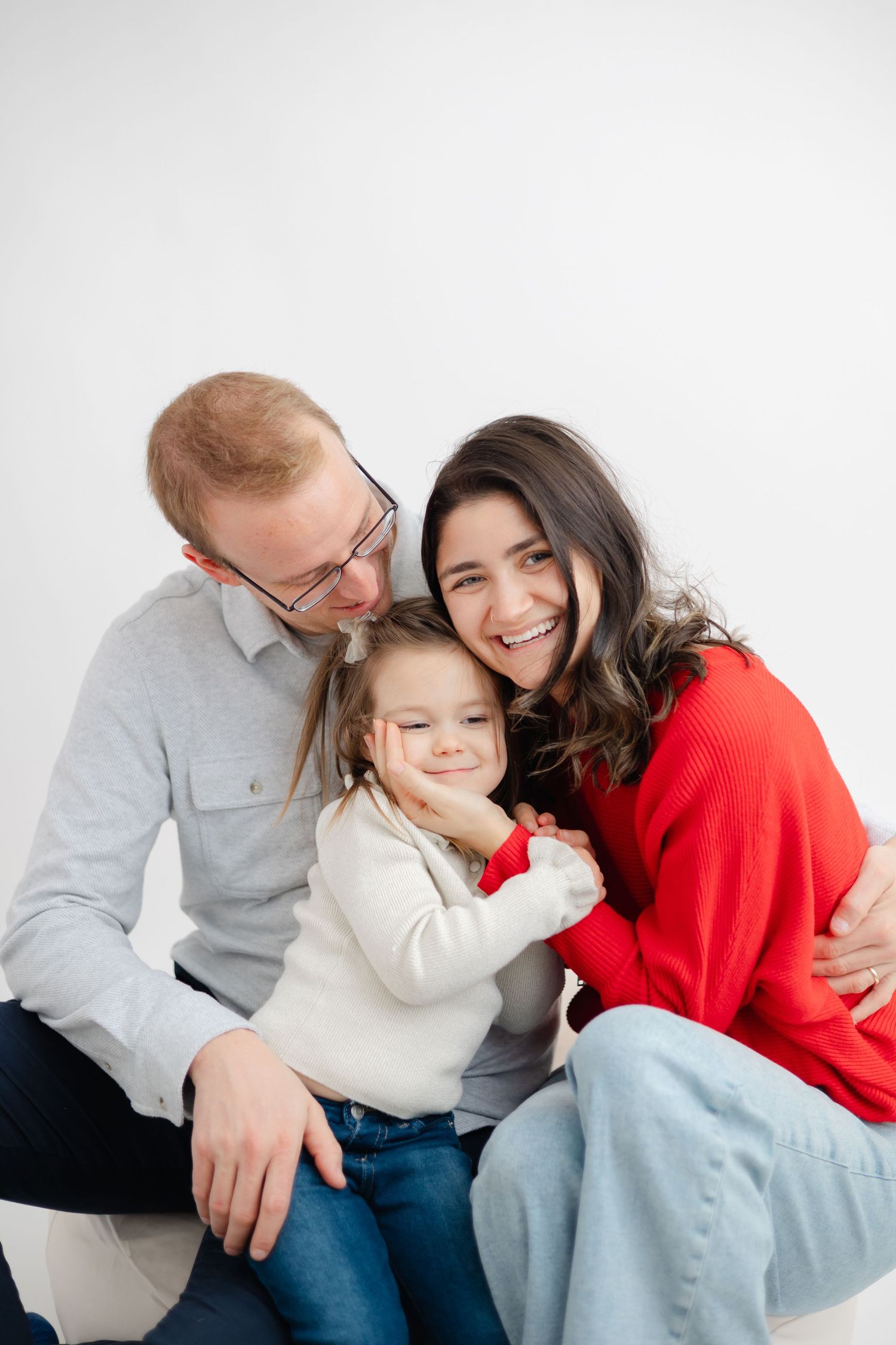 Family of three sharing joyful moments together in a bright studio setting wearing red and light colored clothing.