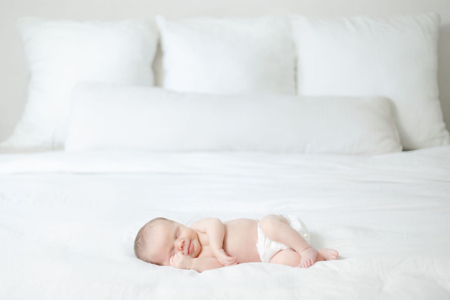 A sleeping newborn baby lies peacefully on white bedding surrounded by soft pillows.