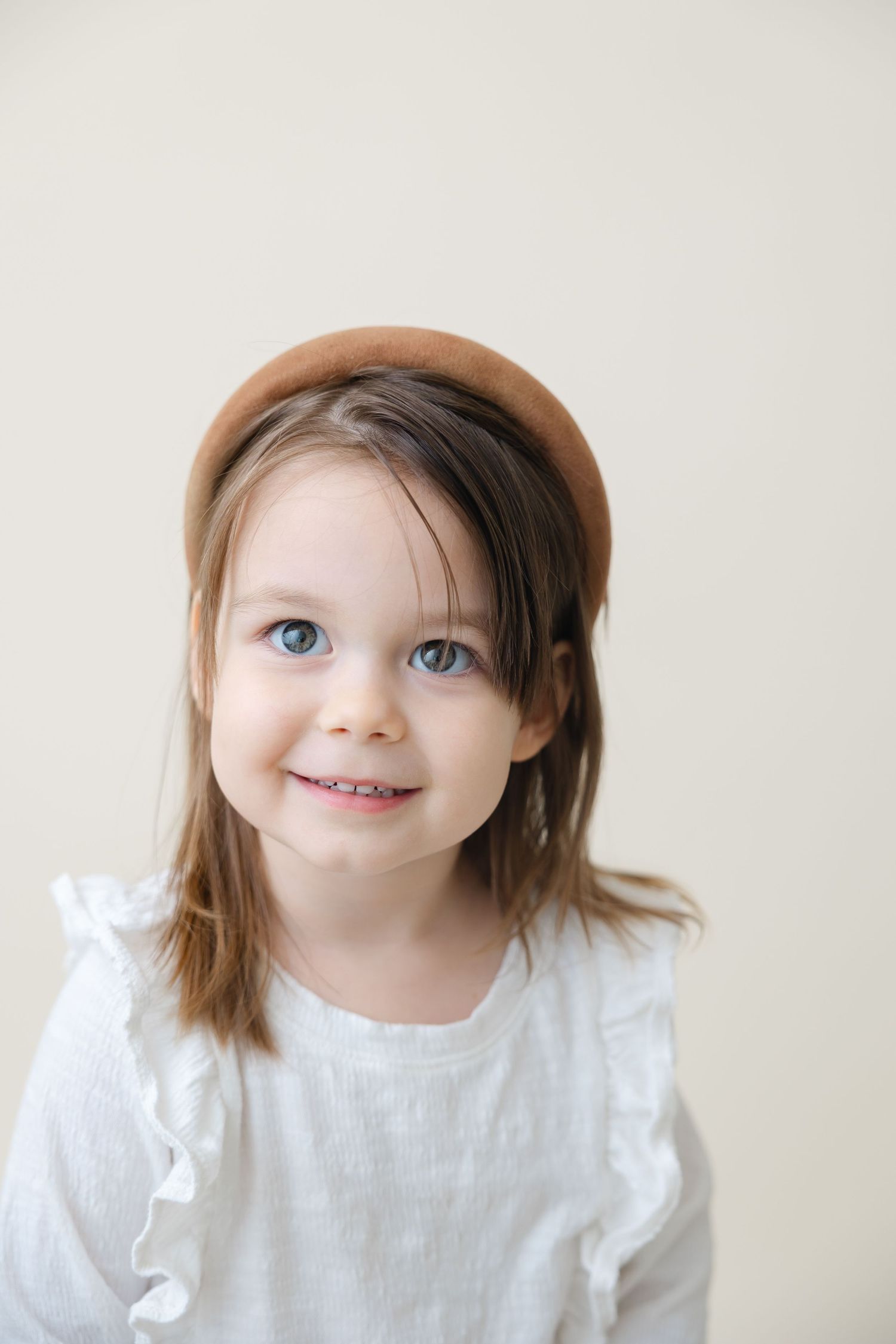Young child wearing a white linen outfit smiles warmly against a light neutral background.