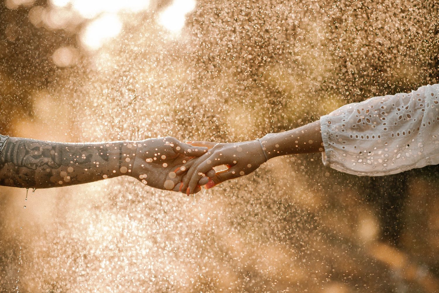 Close-up of hands reaching for each other through water droplets at sunset.