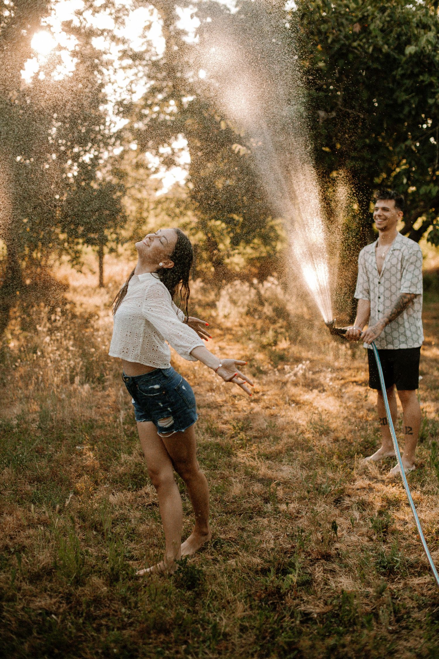 Playful couple enjoys water spray in golden sunlit garden setting.