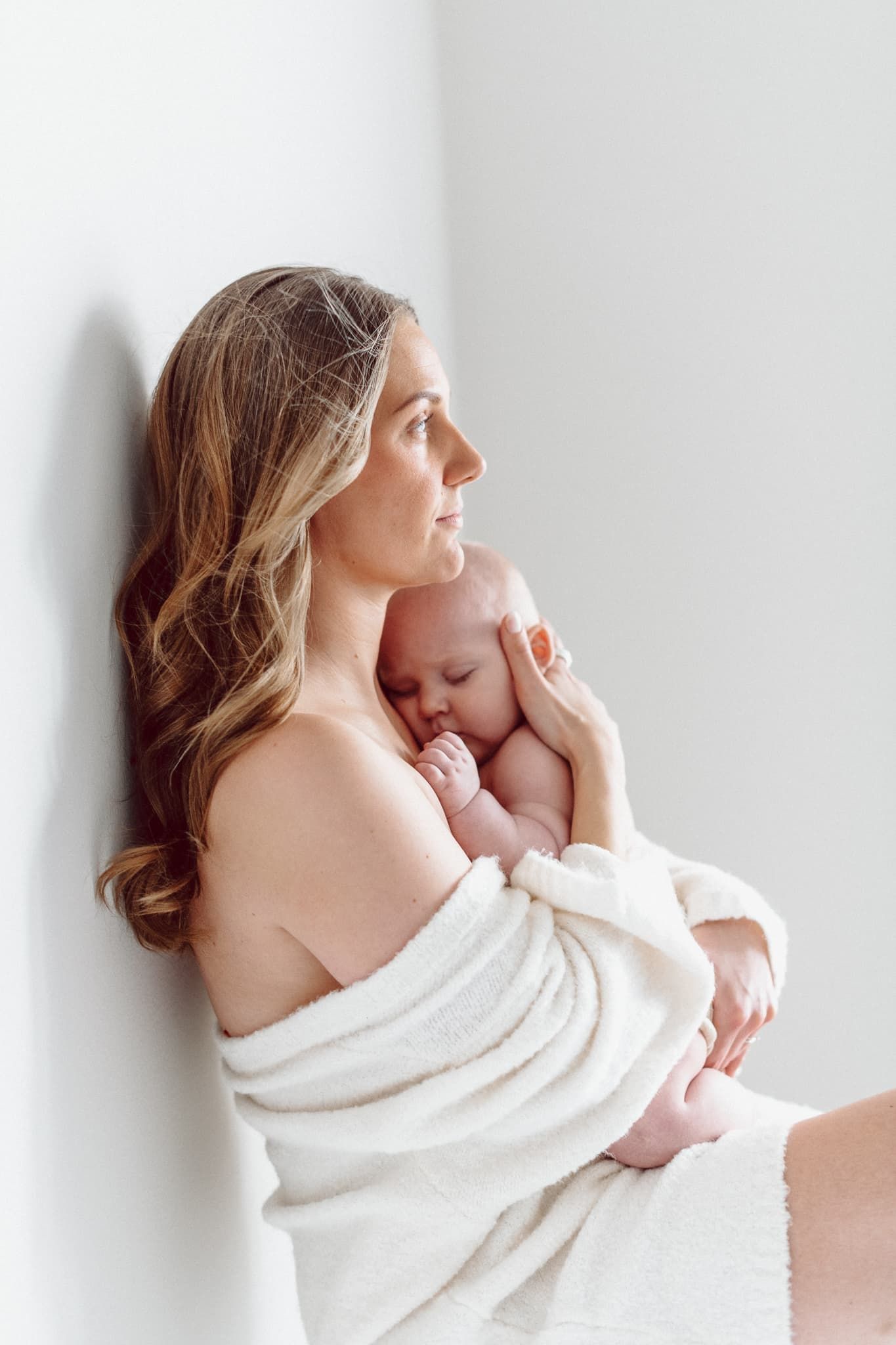 Mamma med vit klänning, sitter på golvet i fotostudio i Mullsjö. Hon håller om sin bebis och tittar ut genom ett fönster med solljus.