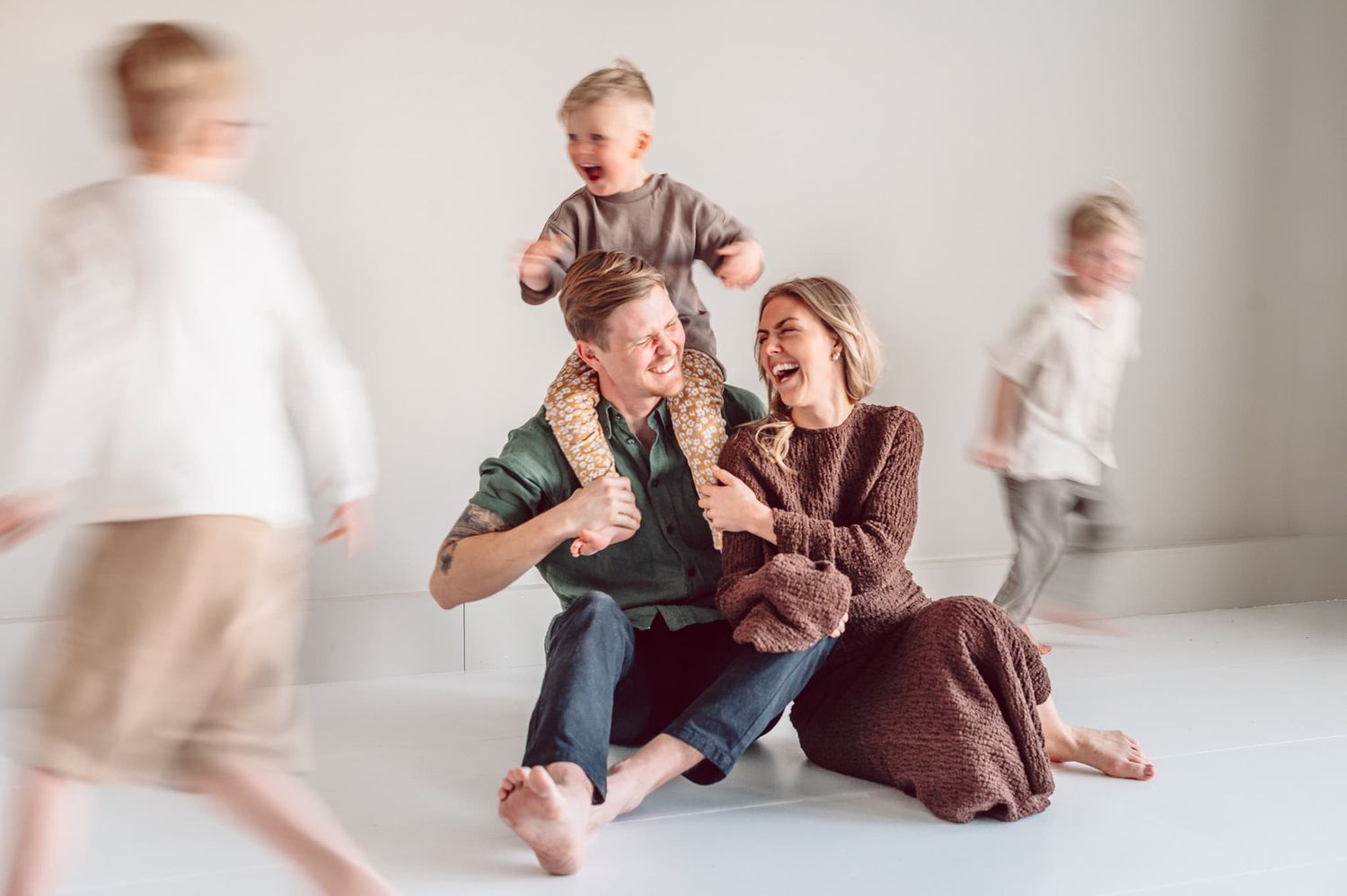 Familj med 3 barn leker i en fotostudio i Jönköping & Mullsjö. Föräldrarna skrattar och barnen springer runt dom. Mamman har brun klänning.