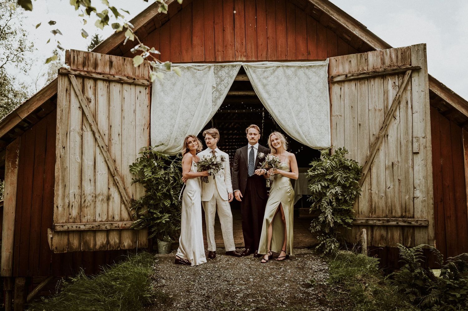 Brudpar med brudfölje står i öppningen av en lada där dom ska ha sin bröllopsfest. Öppningen har bohemiska gardiner och gröna blad.