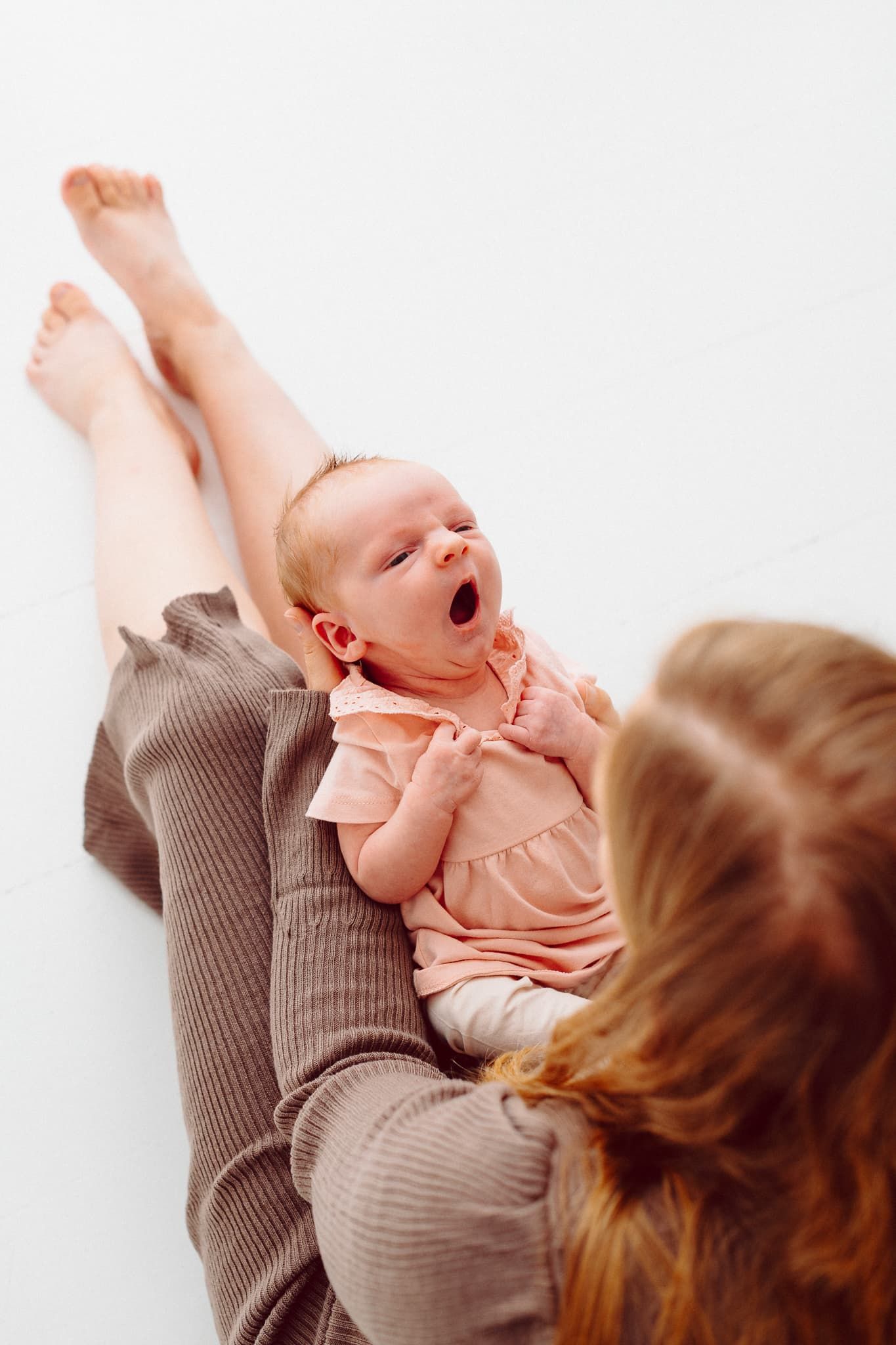 Nyfödd flicka ligger i sin mamma knä och gäspar. Mamman har brun klänning och bebisen rosa klänning.