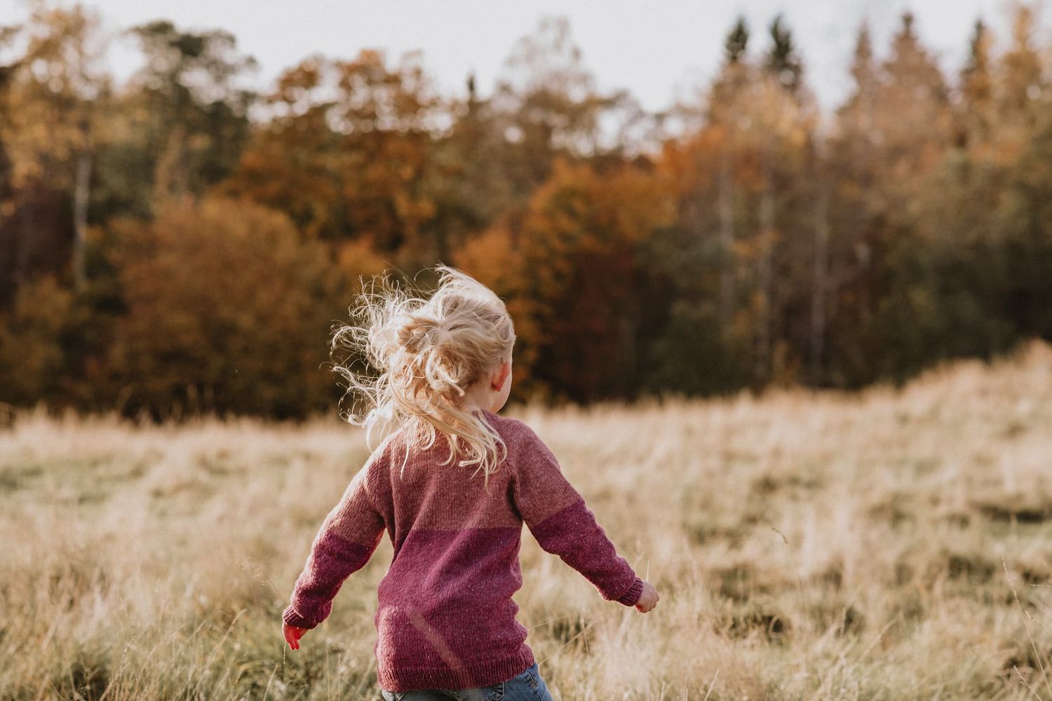 Flicka i stickad rosa tröja springer på en äng. Det blonda håret fladdrar i vinden.