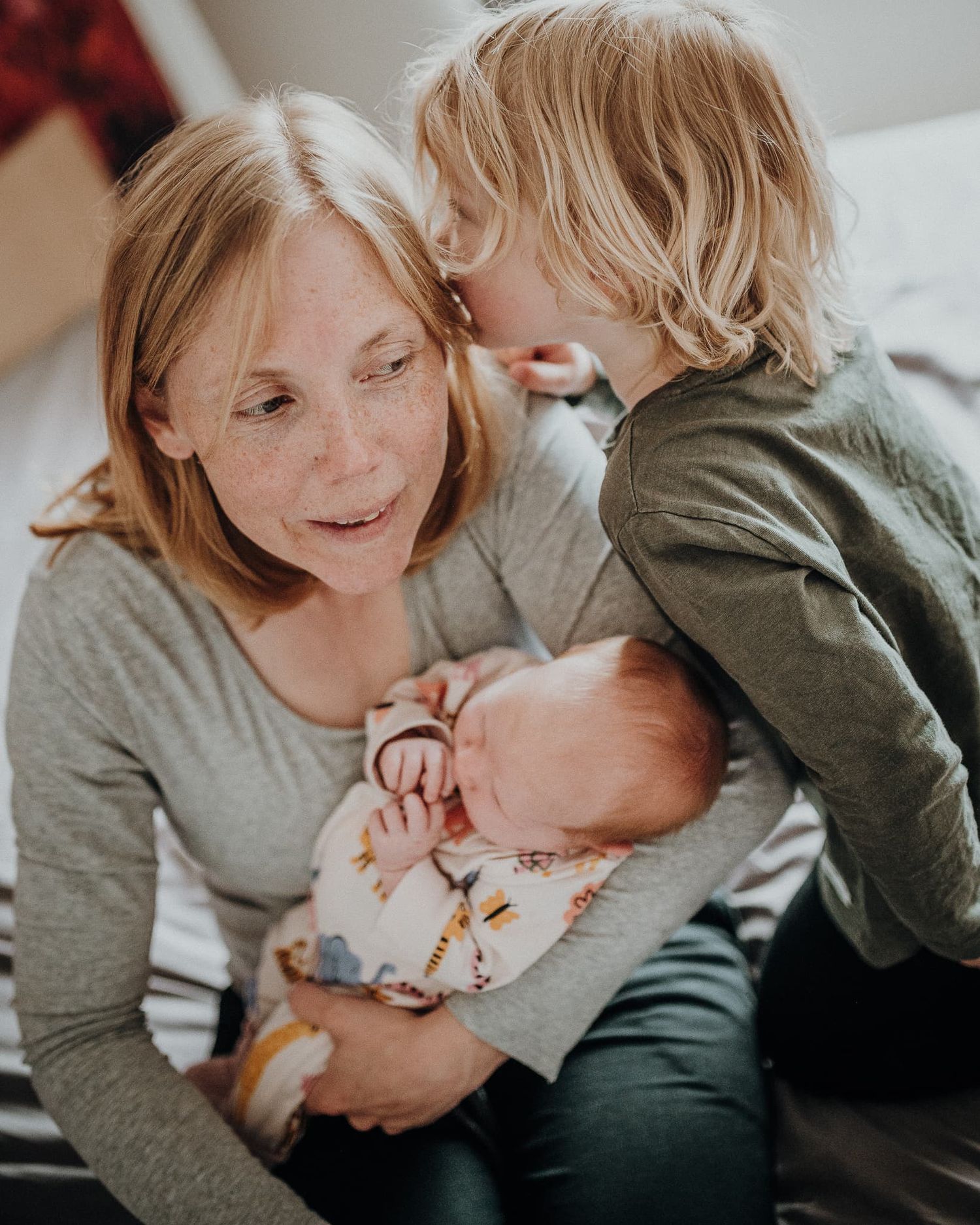 Mammasitter på sängkant med en bebis i famnen. Hennes äldre son viskar i hennes öra.