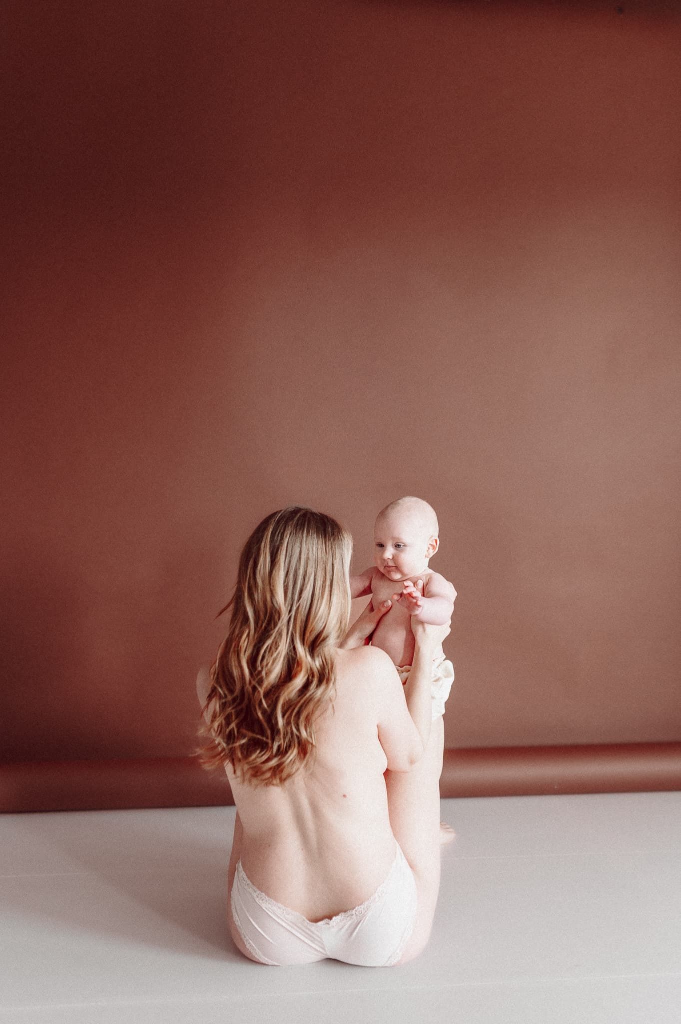 Mamma sitter på golvet i fotostudio i Mullsjö. Hon håller sin bebis. Hon har bar hud och underbyxor.