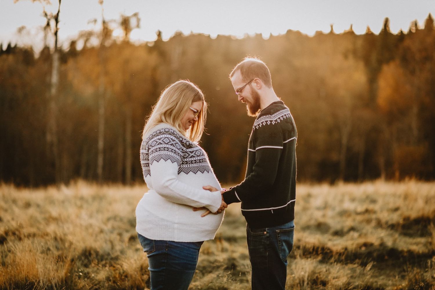Gravid kvinna och hennes man håller om gravidmagen. Dom står på en äng på hösten och det är ett varmt kvällsljus i bilden.