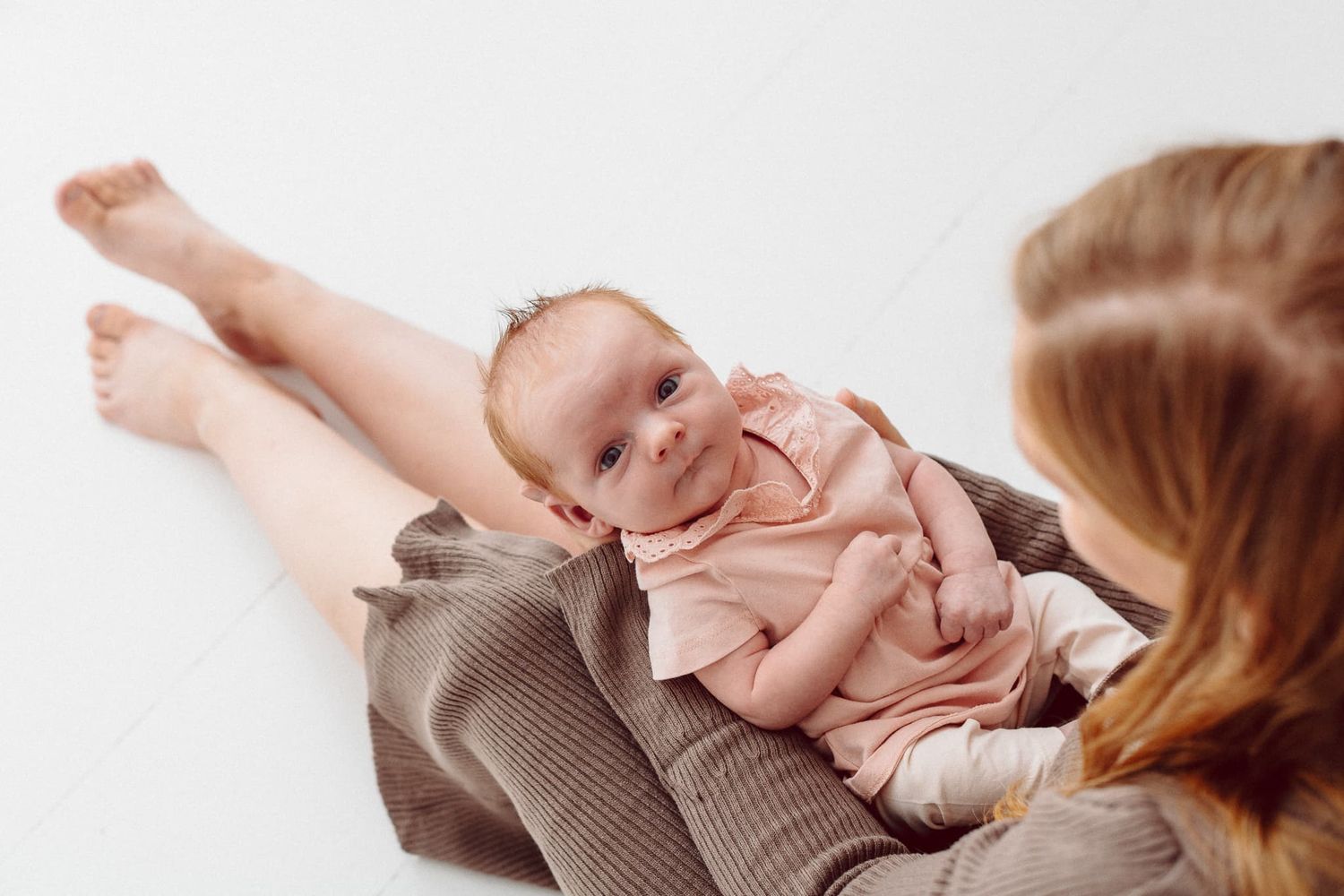Nyfödd bebis ligger i sin mammas knä. Mamma sitter på ett vitt golv i fototudio hos Fotograf Veronica Eng. Mammahar en brun klänning.