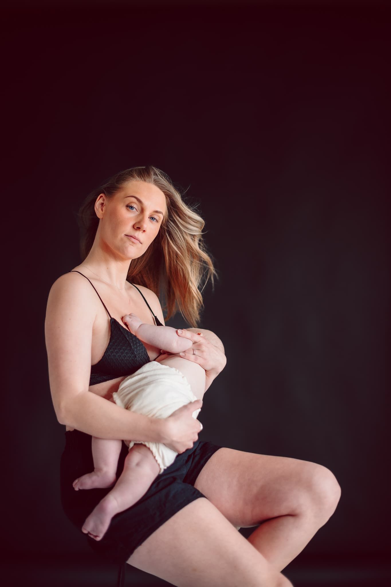 Mamma sitter i fotostudio i Mullsjö. Bakgrunden är svart och hon har svarta underkläder. Hon håller sin bebis och håret fladdrar.