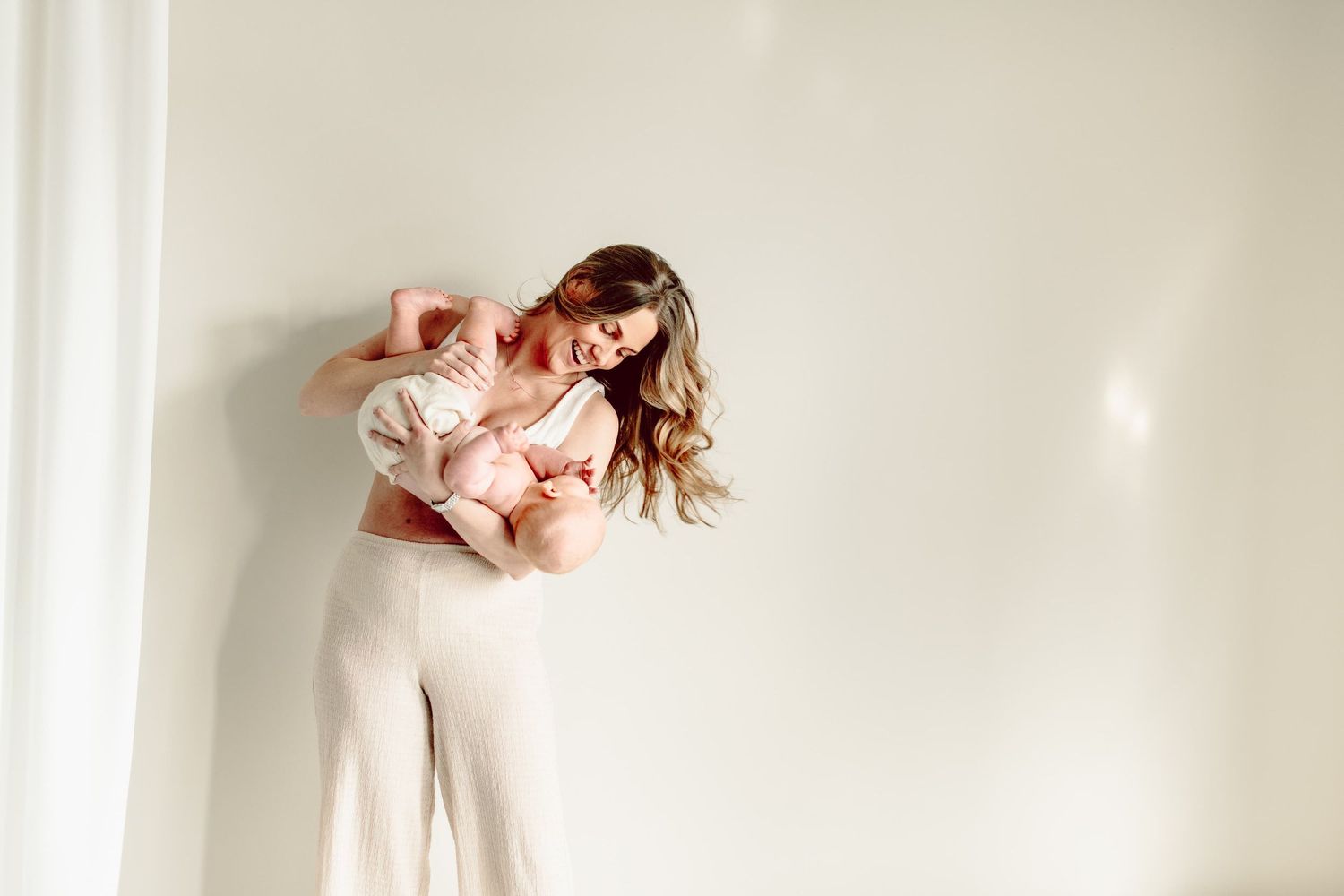 Moderskapsfotografering i Studio. Mamman har vita byxor och busar med sim lilla bebis som har vita blöjbyxor. Studion är ljus.