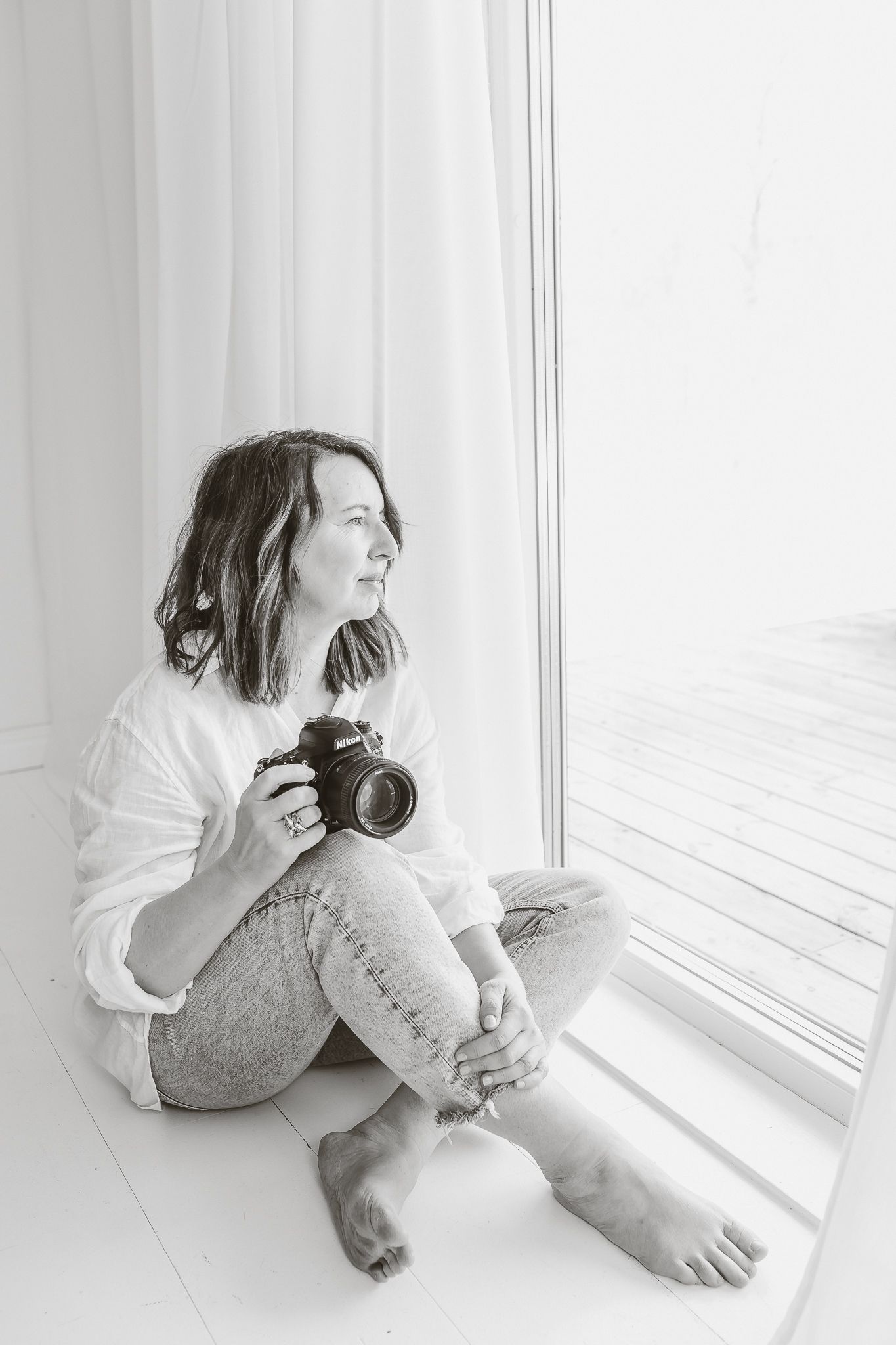 Veronica Eng sitter på golvet i sin fotostudio.Hon har en vit skjorta och jeans.Hon håller i sin Nikon kamera och ser ut genom ett fönster.