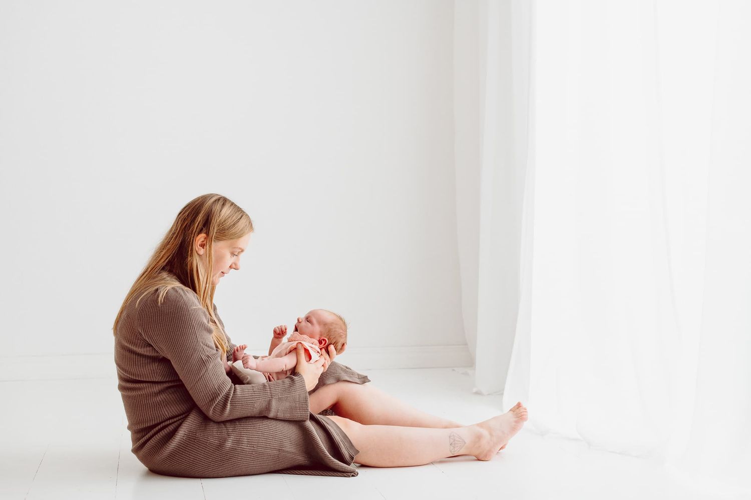 Mamma sitter på golvet i en vit studio. Hon har sin nyfödda bebis i famnen. Hon har brun klänning och bebisen rosa klänning.