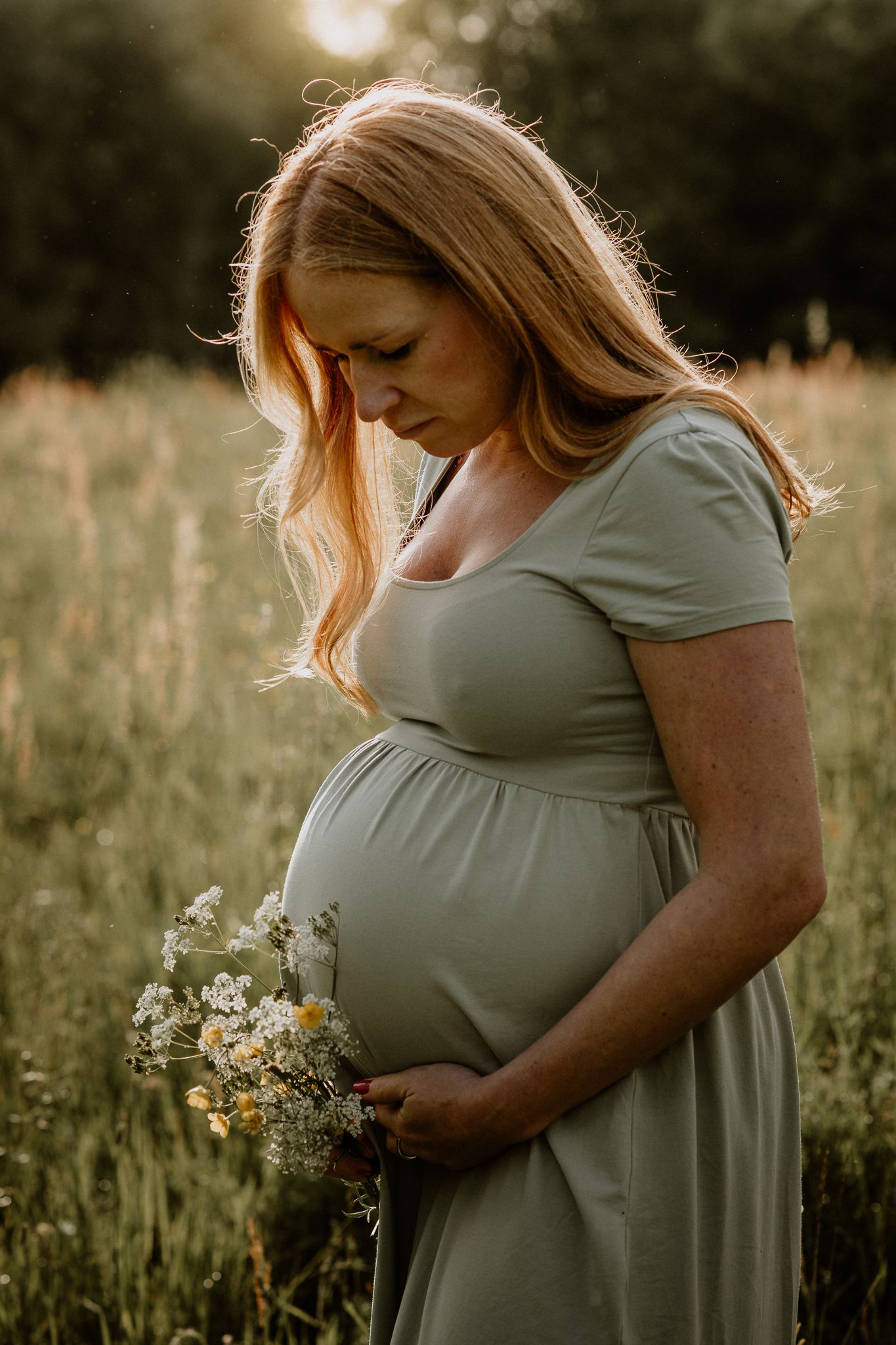 Gravid kvinna i grön klänning står i motljus på en äng. Hon har blommor i handen och tittar ner på sin gravidmage.